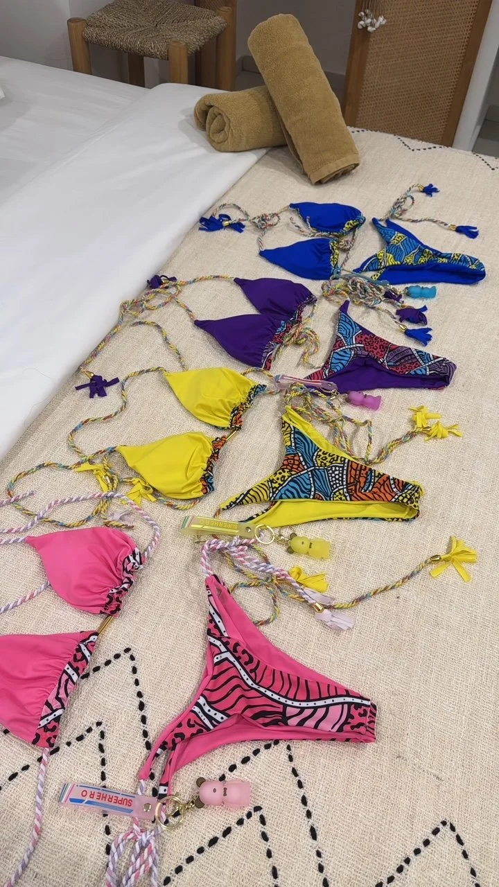 Colorful bikini swimsuits with matching tie strings and small plush keychains arranged on a beige surface next to a bed with rolled towels and chairs in the background.