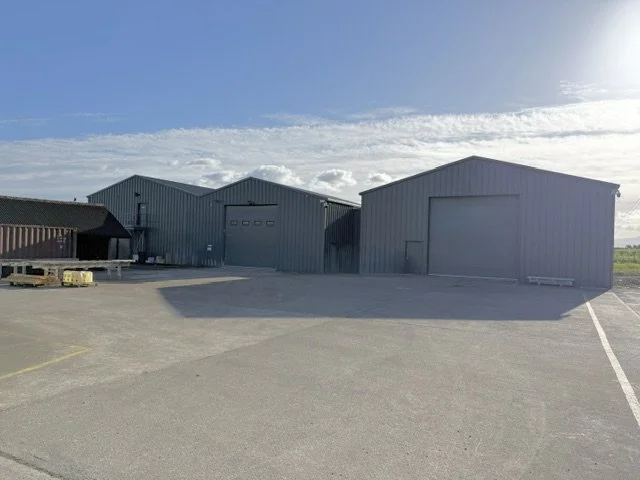 Three large metal storage or warehouse buildings in an open lot with a clear blue sky and some clouds above.