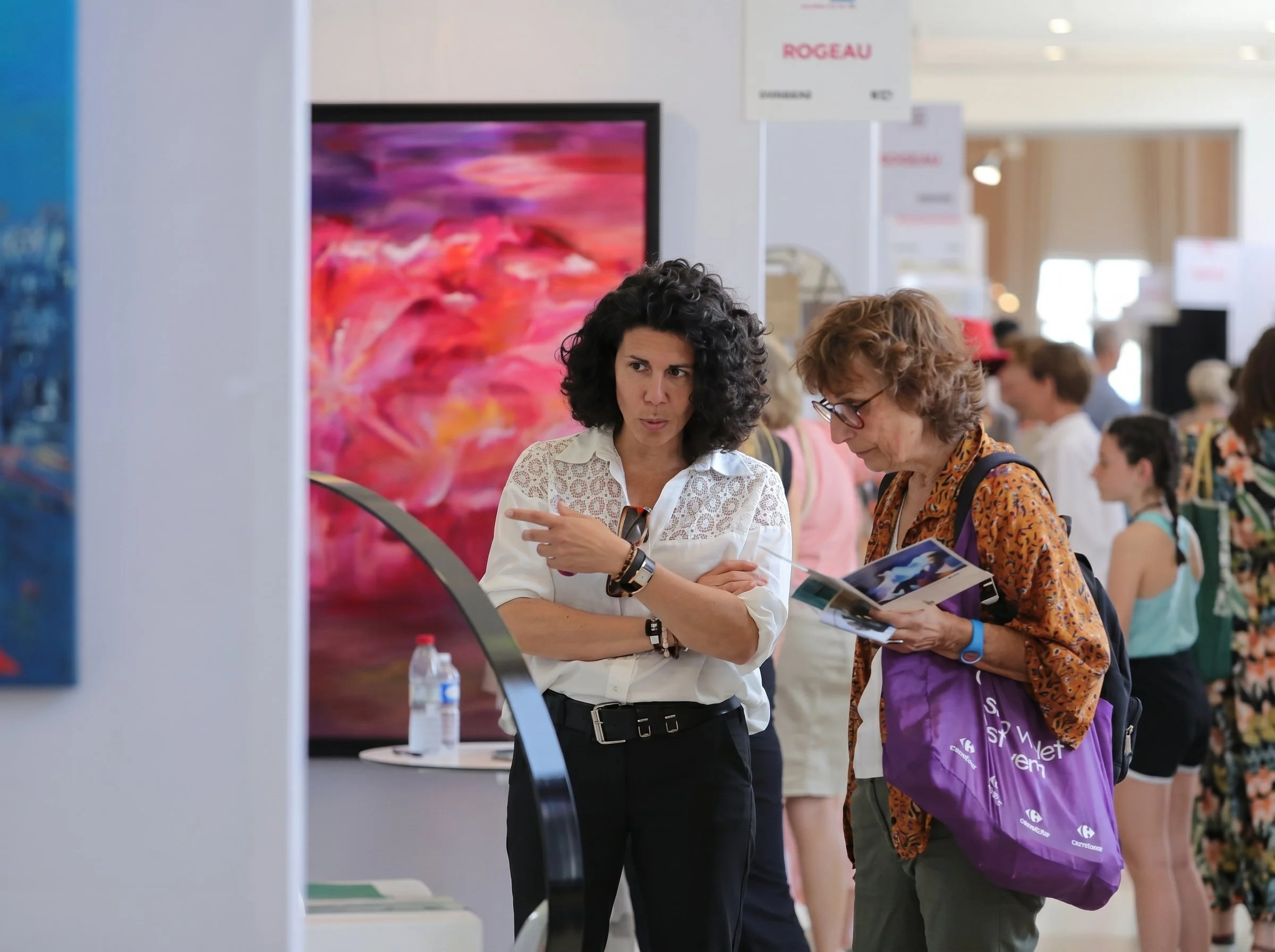 Deux femmes discutent à une foire ou un salon d'exposition, entourées d'autres visiteurs et d'œuvres d'art ou expositions en arrière-plan.