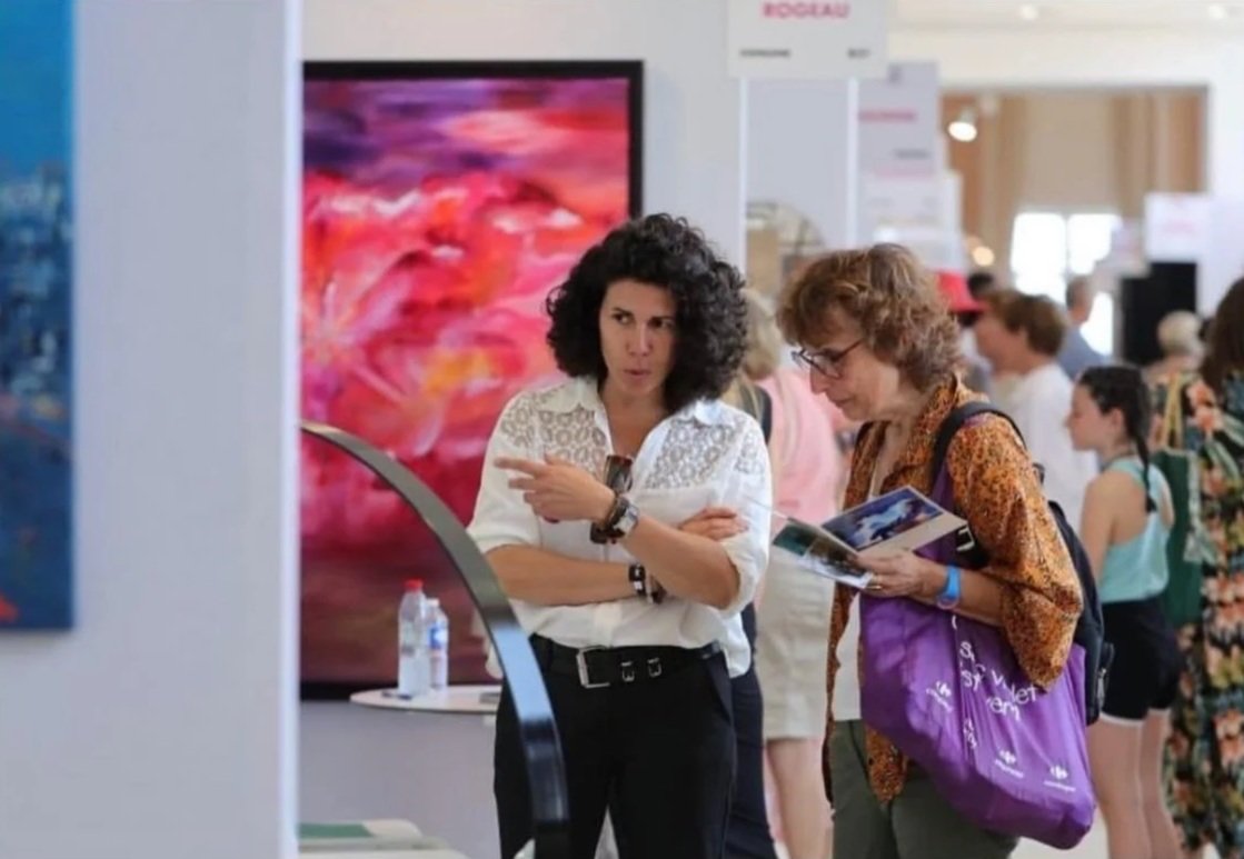 Deux femmes discutent devant une œuvre d'art abstraite lors d'une exposition, en lisant un dépliant.