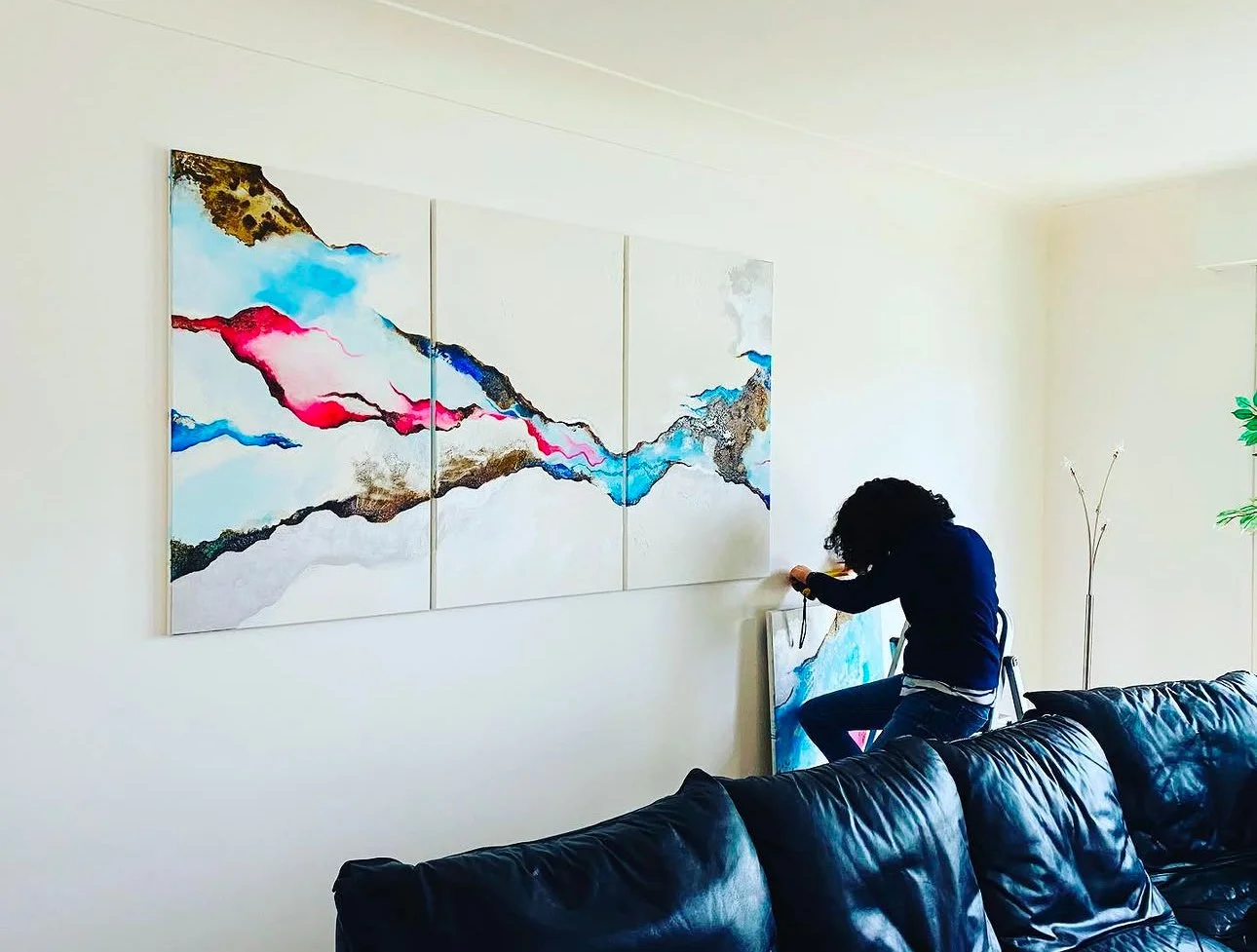 Une personne assise sur une chaise, en train de créer ou d'ajuster une peinture abstraite dans un salon, avec un grand tableau coloré accroché au mur.