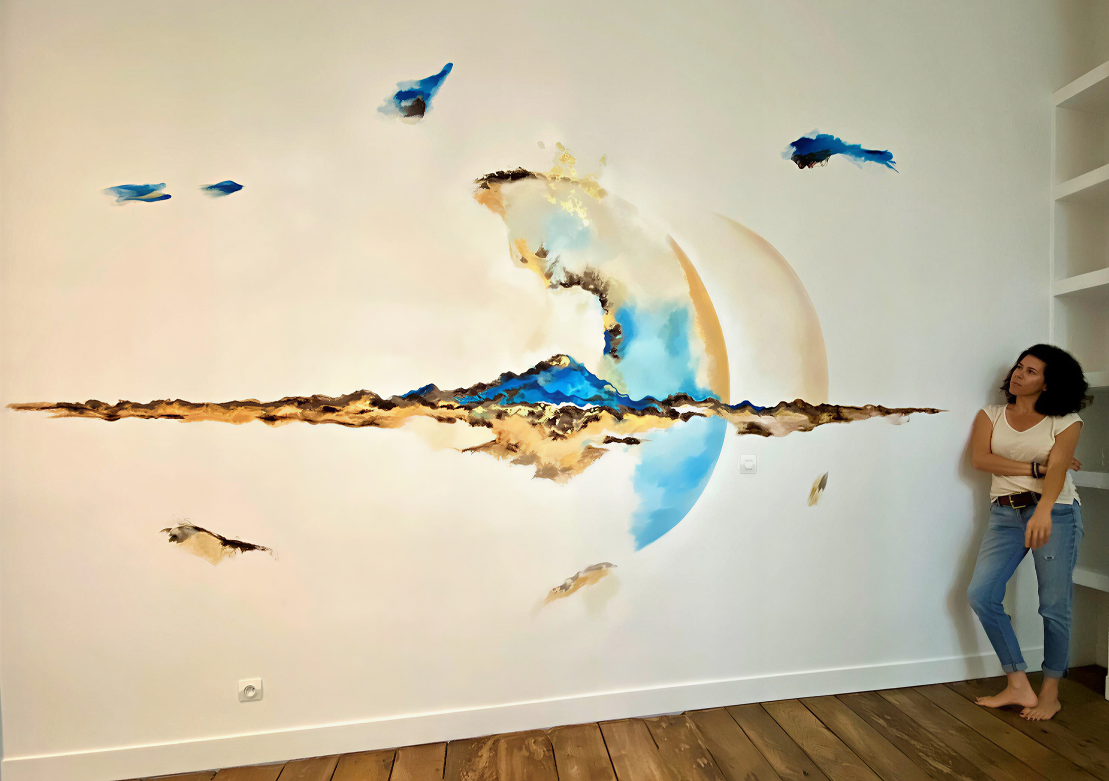 Une femme debout contre un mur blanc, à côté d'une grande œuvre d'art abstraite représentant une explosion ou une éruption avec des couleurs chaudes et froides, notamment du bleu, blanc, orange et noir.
