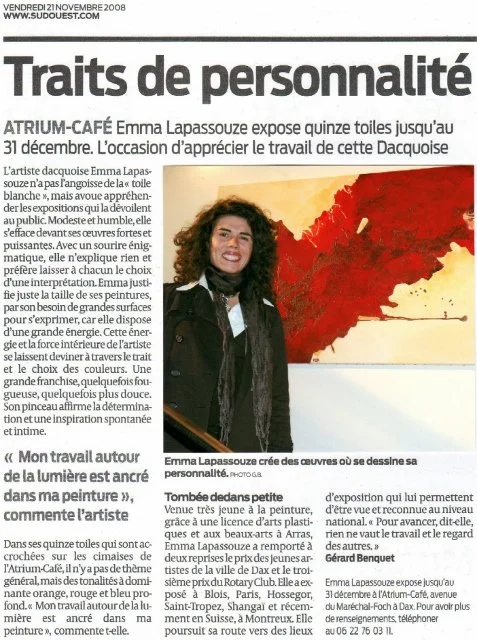 Une femme, Emma Lapassouze, souriante, devant une peinture de fleurs aux couleurs rouge, orange et jaune, dans une galerie ou un espace d'exposition.