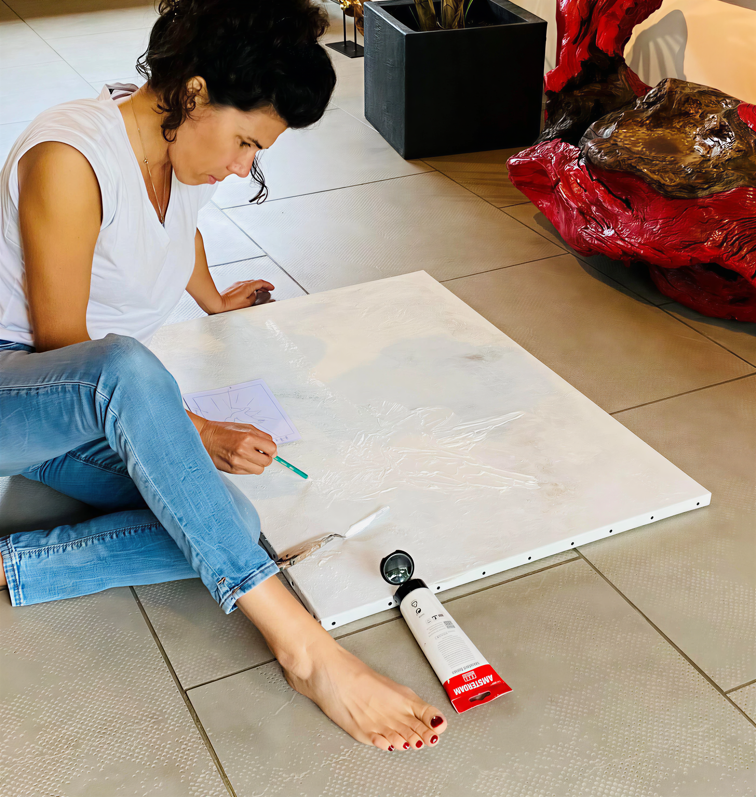 Une femme allongée sur un sol en céramique, tenant un stylo, avec une grande masse en plastique rouge à côté d'elle et un tube blanc à ses pieds. Elle semble faire un dessin ou écrire.