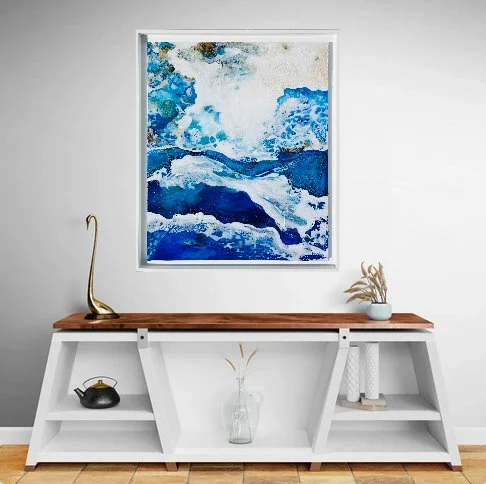 Peinture abstraite représentant des vagues de mer en nuances de bleu, blanc et beige, encadrée et suspendue sur un mur blanc.