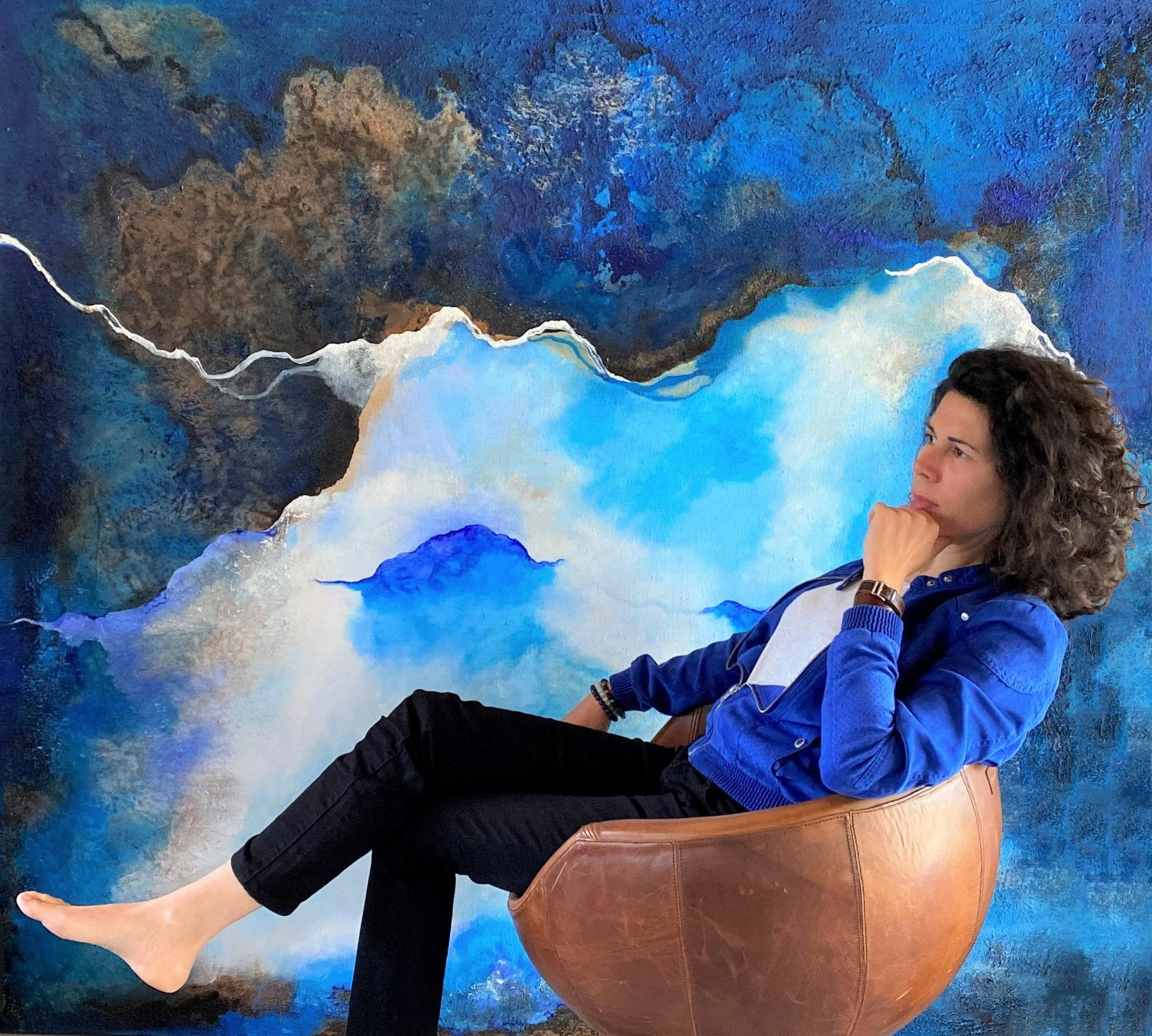 Une femme assise dans un fauteuil en cuir marron, en face d'un tableau abstrait avec des tons de bleu, blanc, beige et noir.