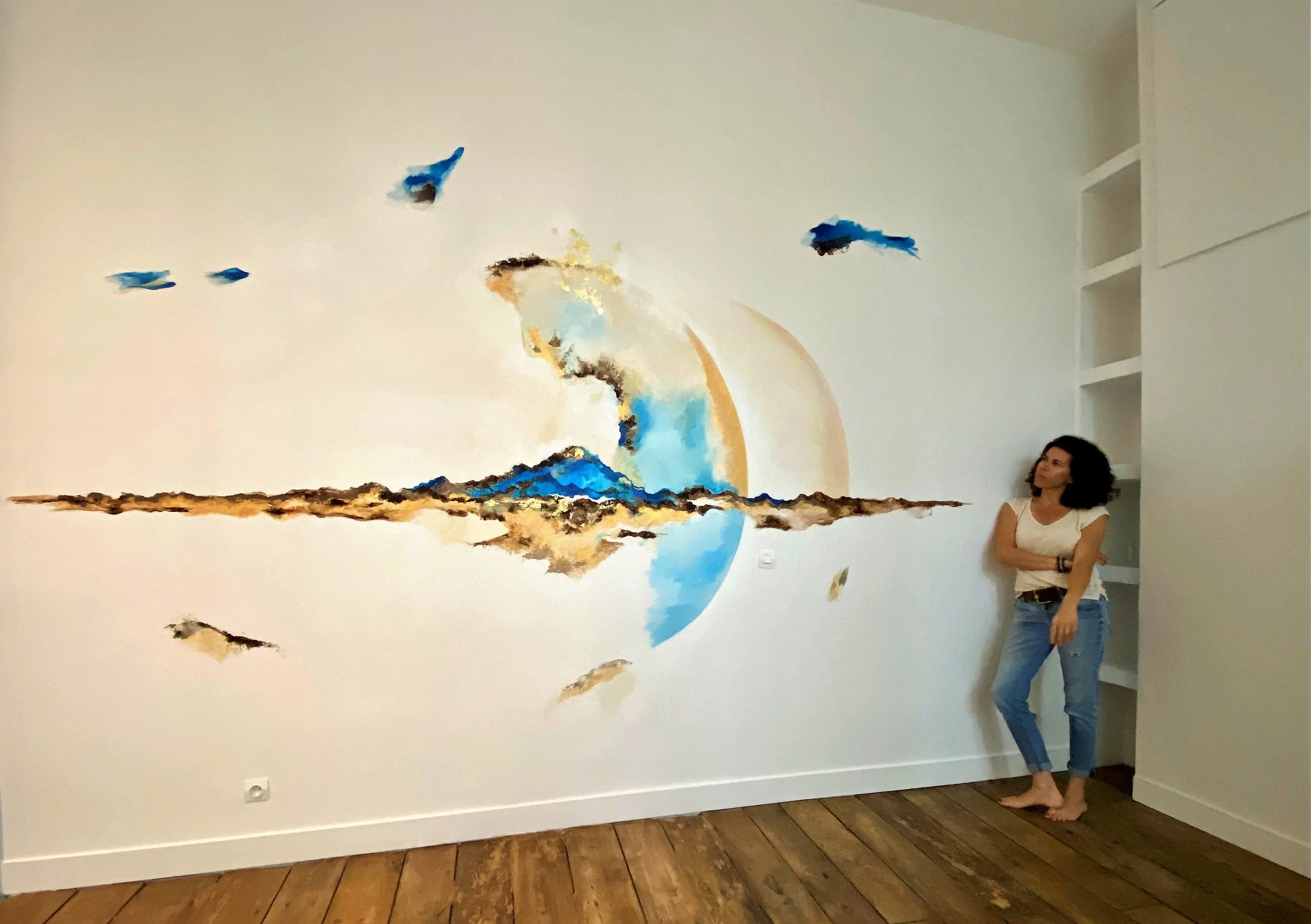 Une femme se tenant à côté d'une grande œuvre d'art abstraite sur un mur blanc, dans une pièce avec un sol en bois.