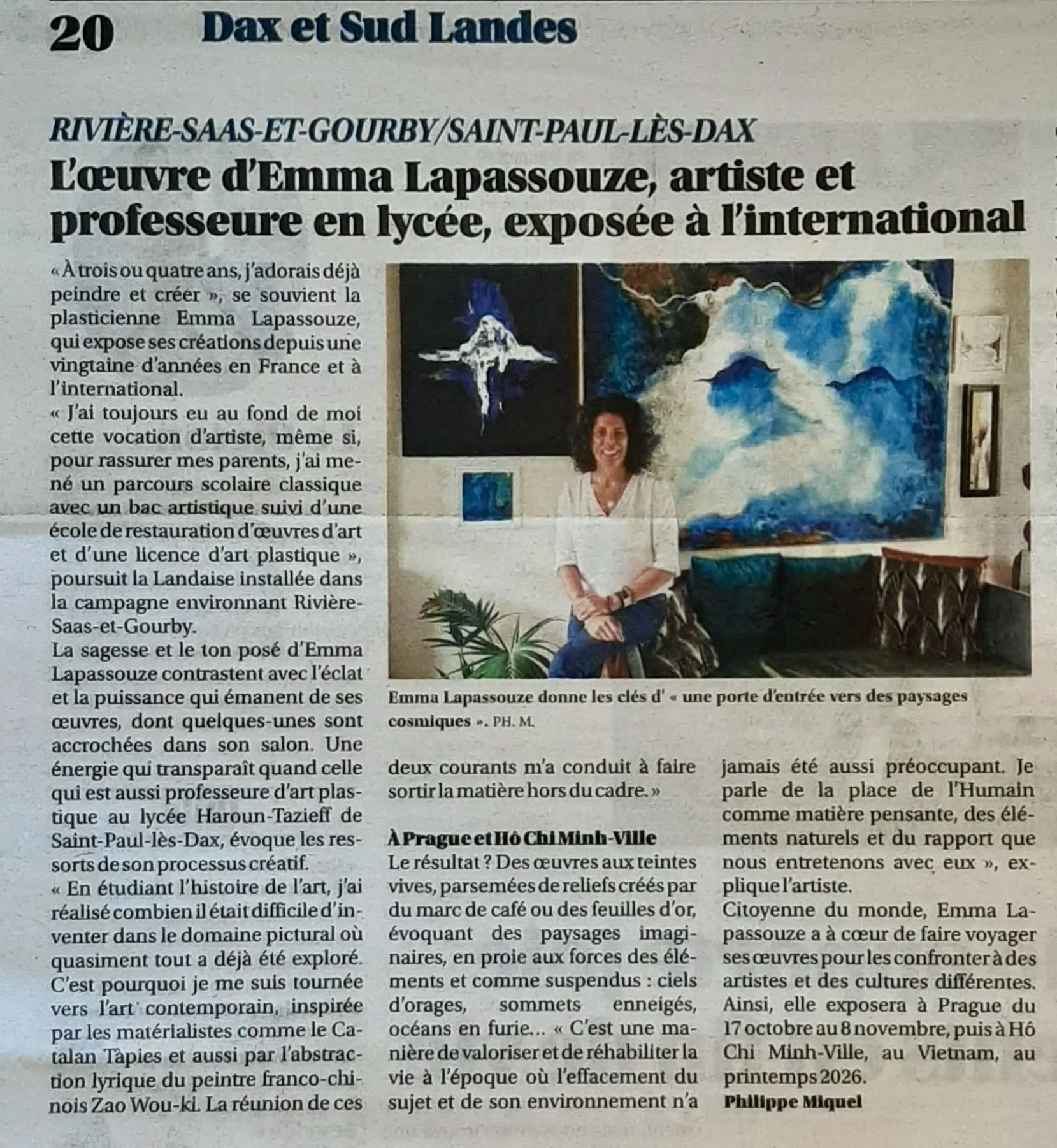 Photographie d'une femme souriante assise dans un salon avec des œuvres d'art abstrait en toile derrière elle, dont un grand tableau bleu et blanc, et d'autres œuvres plus petites. La pièce comporte aussi des meubles en bois et plusieurs plantes.