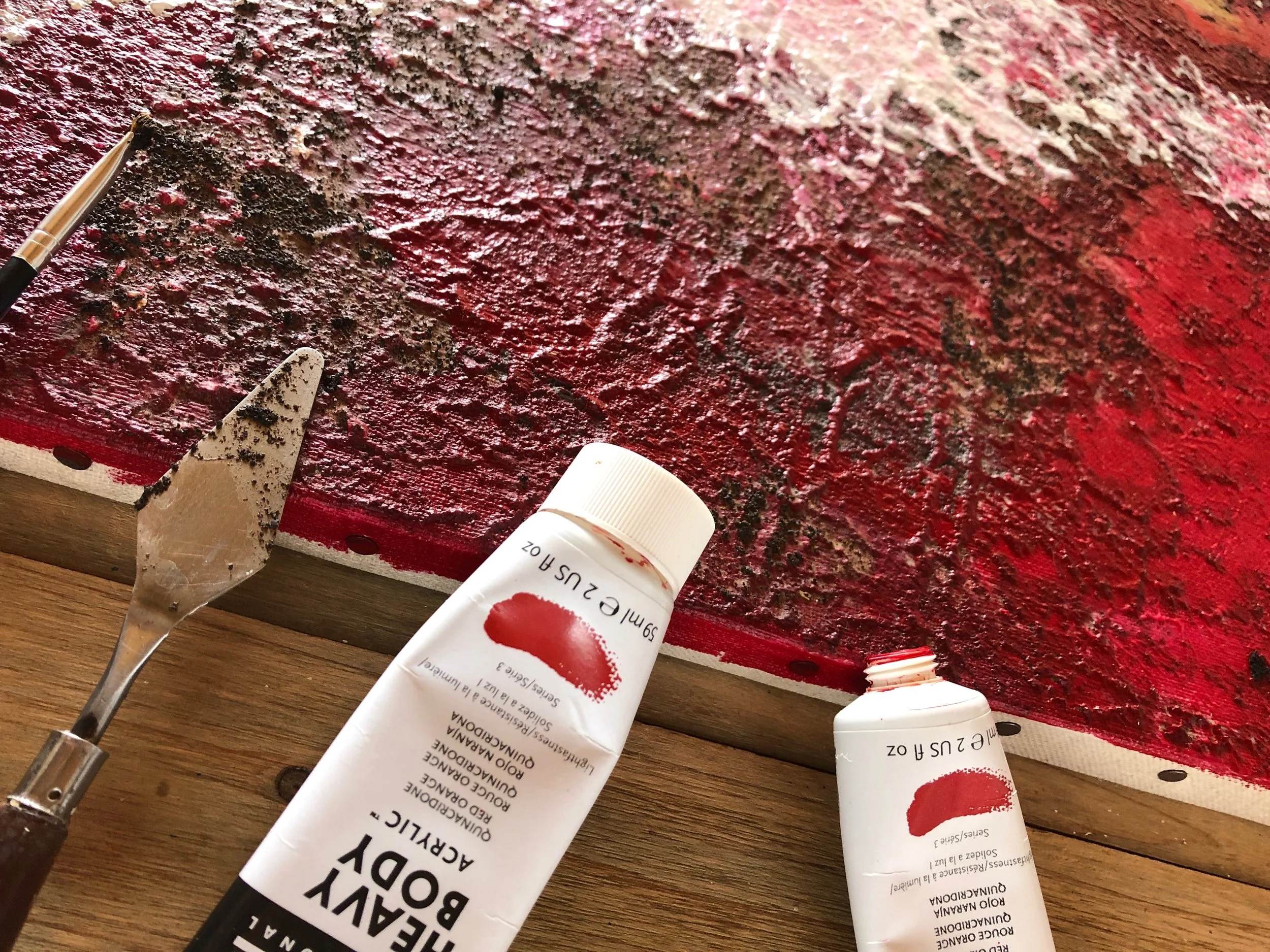 Une toile de peinture rouge en cours de création avec un pinceau, deux tubes de peinture blanche et rouge, posés sur une surface en bois.