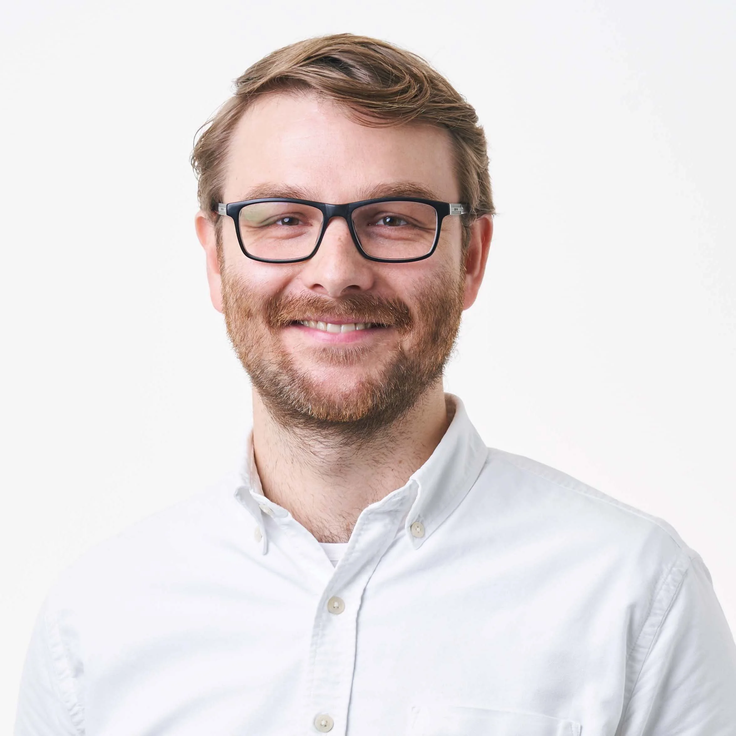 Close-up of a smiling man with glasses and reddish-brown hair, wearing a white button-up shirt against a plain white background.