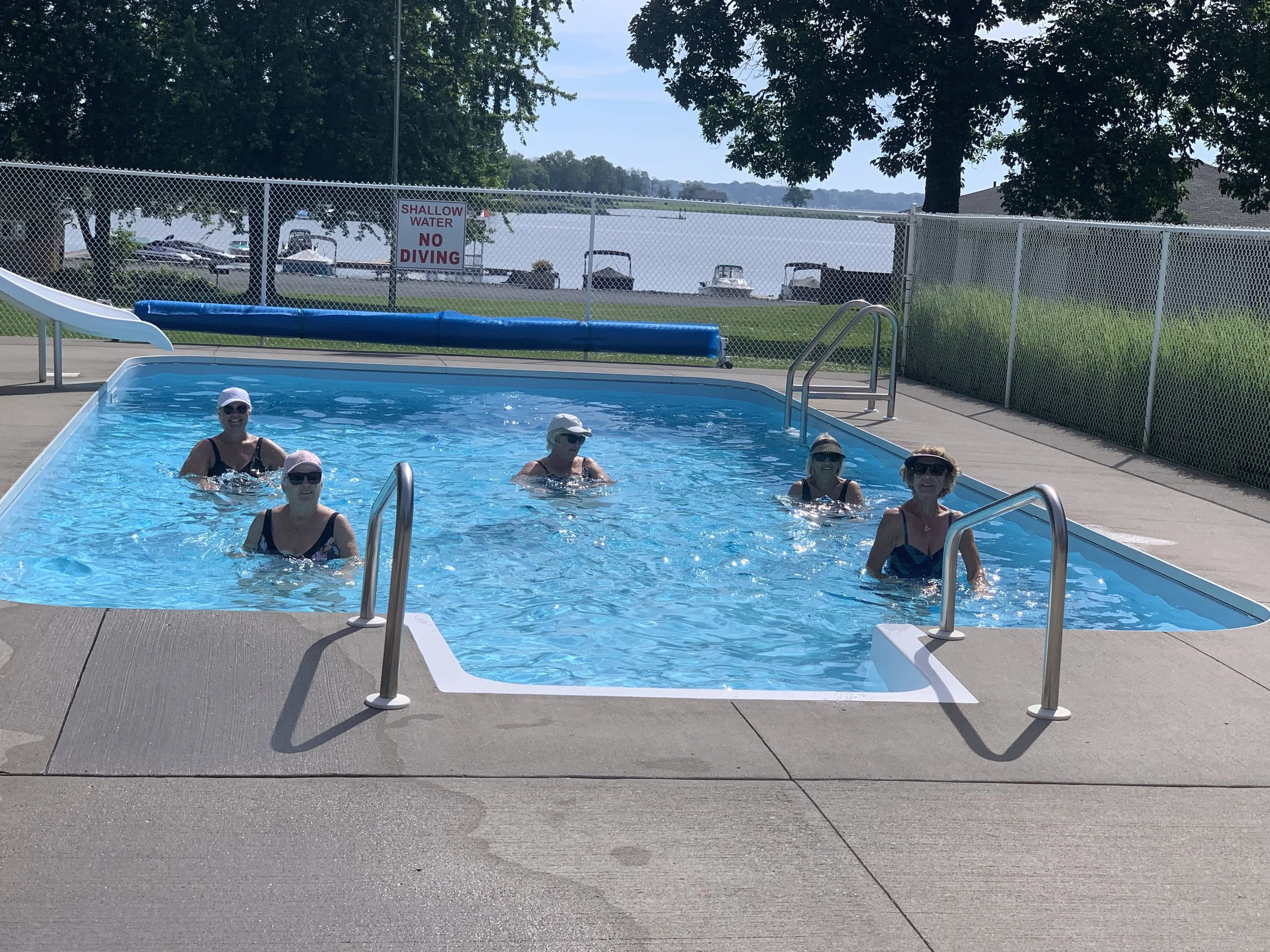 A swimming pool with four women enjoying the water, wearing swimsuits and hats. There is a blue pool float on the pool's edge, and a sign in the background that says 'Shallow Water No Diving'. The pool area is surrounded by a fence, trees, and a view