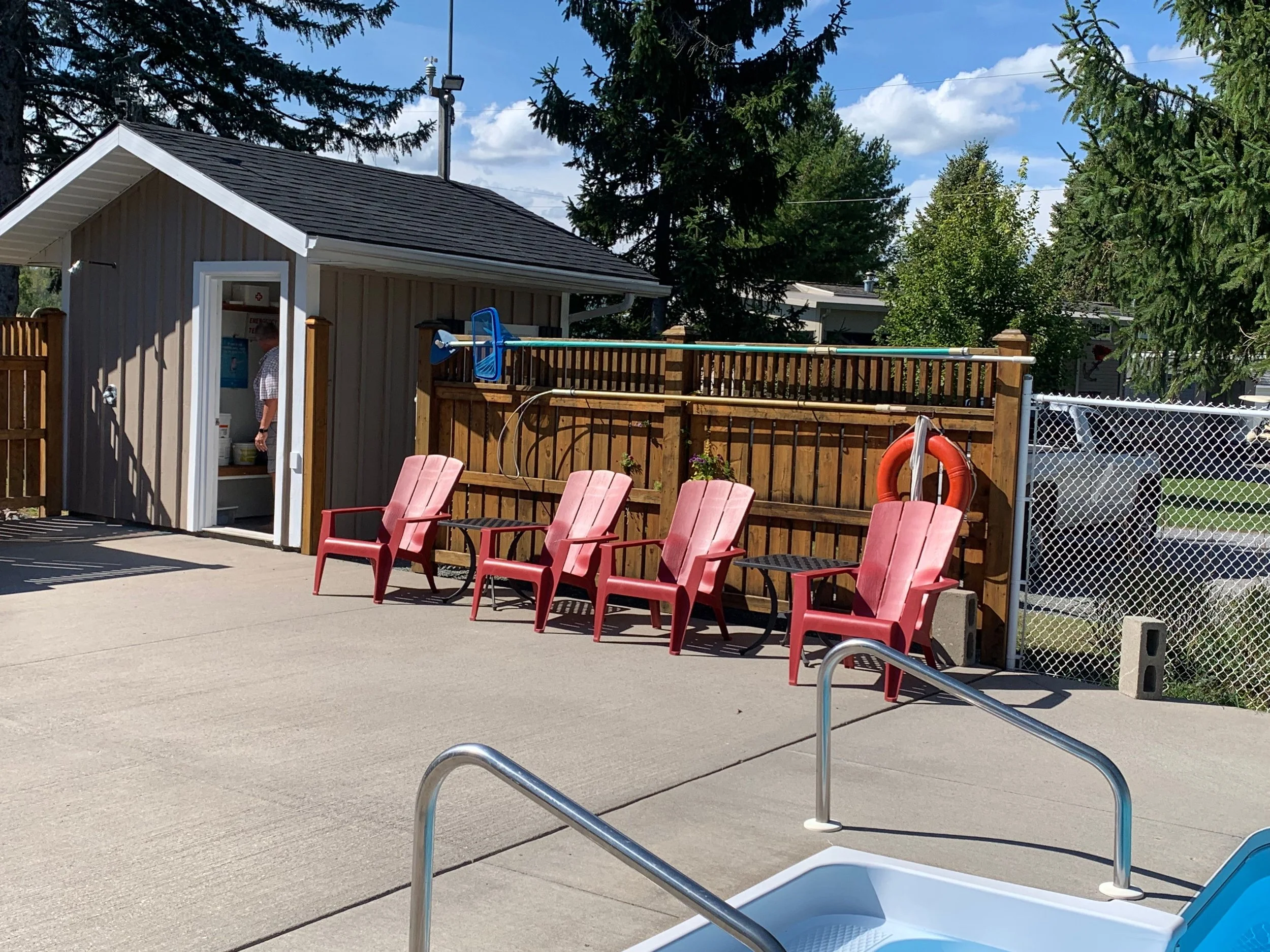 An outdoor pool area with lounge chairs, a fenced-in area, and a small shed. There are trees and blue sky with some clouds in the background.