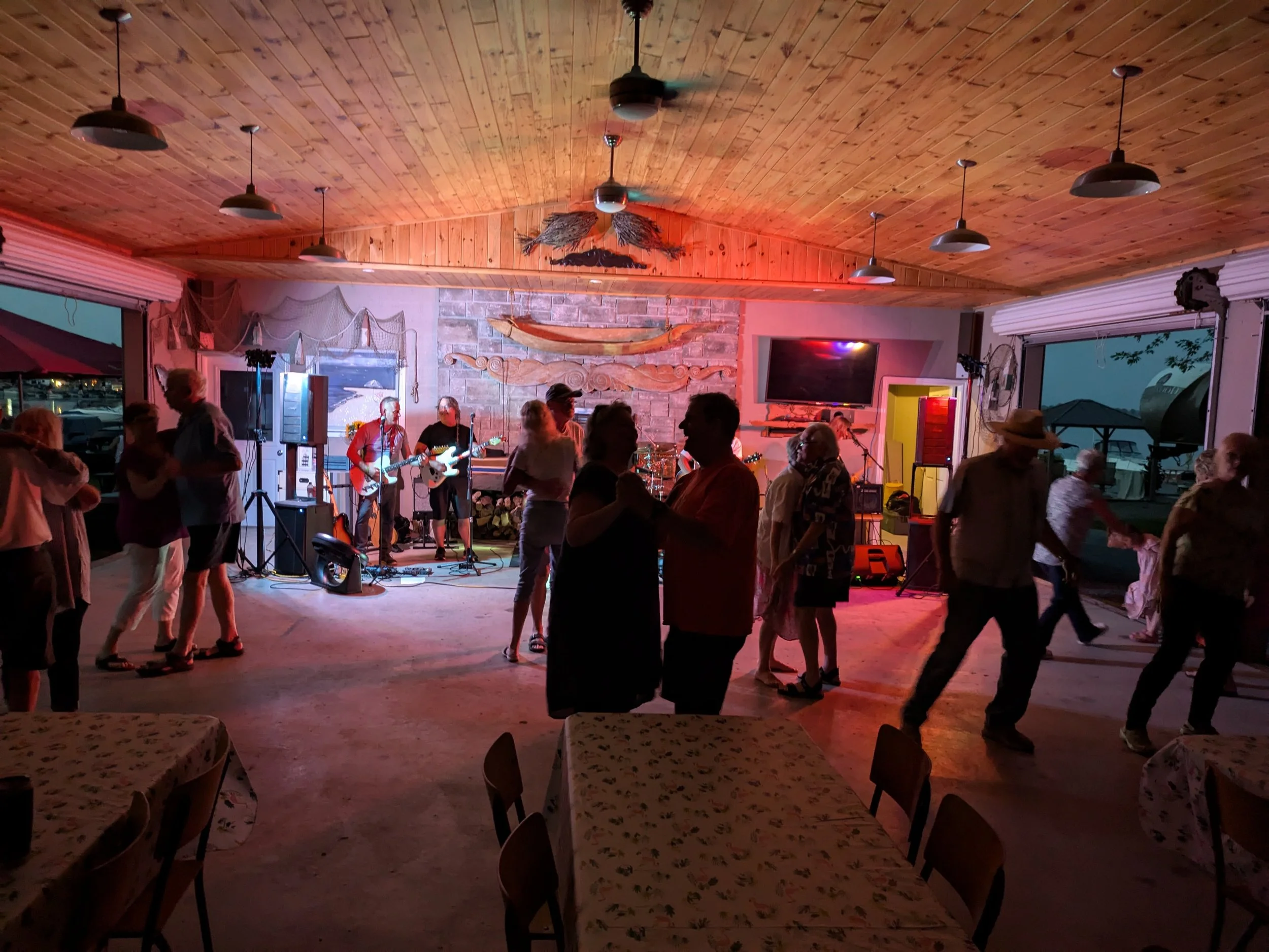 Indoor venue with a wooden ceiling, people dancing, a live band performing on stage, and tables with floral tablecloths.