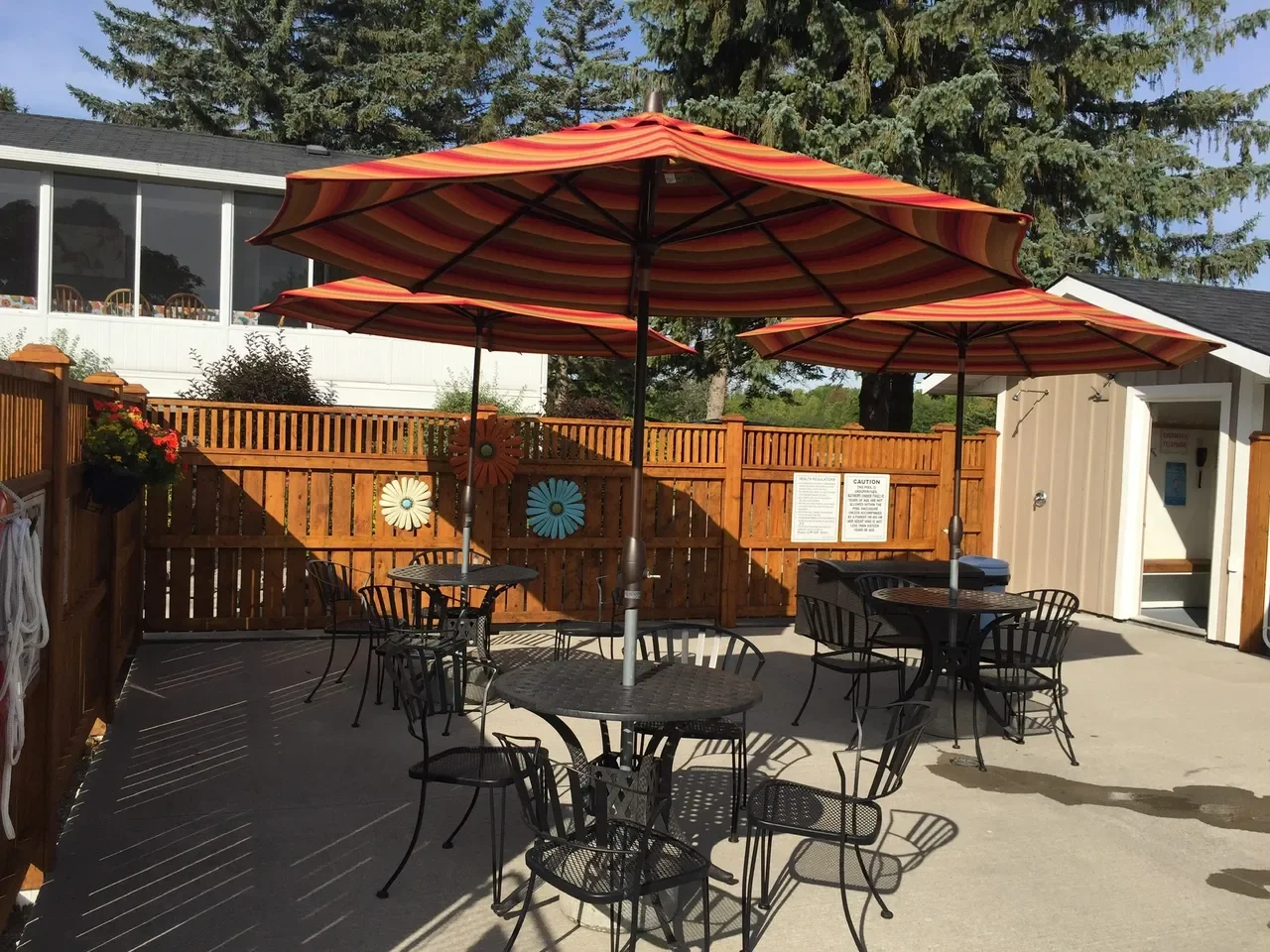 Outdoor patio with black metal tables and chairs, shaded by three large orange and yellow striped umbrellas. A wooden fence with decorative flower art and a building with a white and gray exterior are visible in the background. Shadows from the umbre