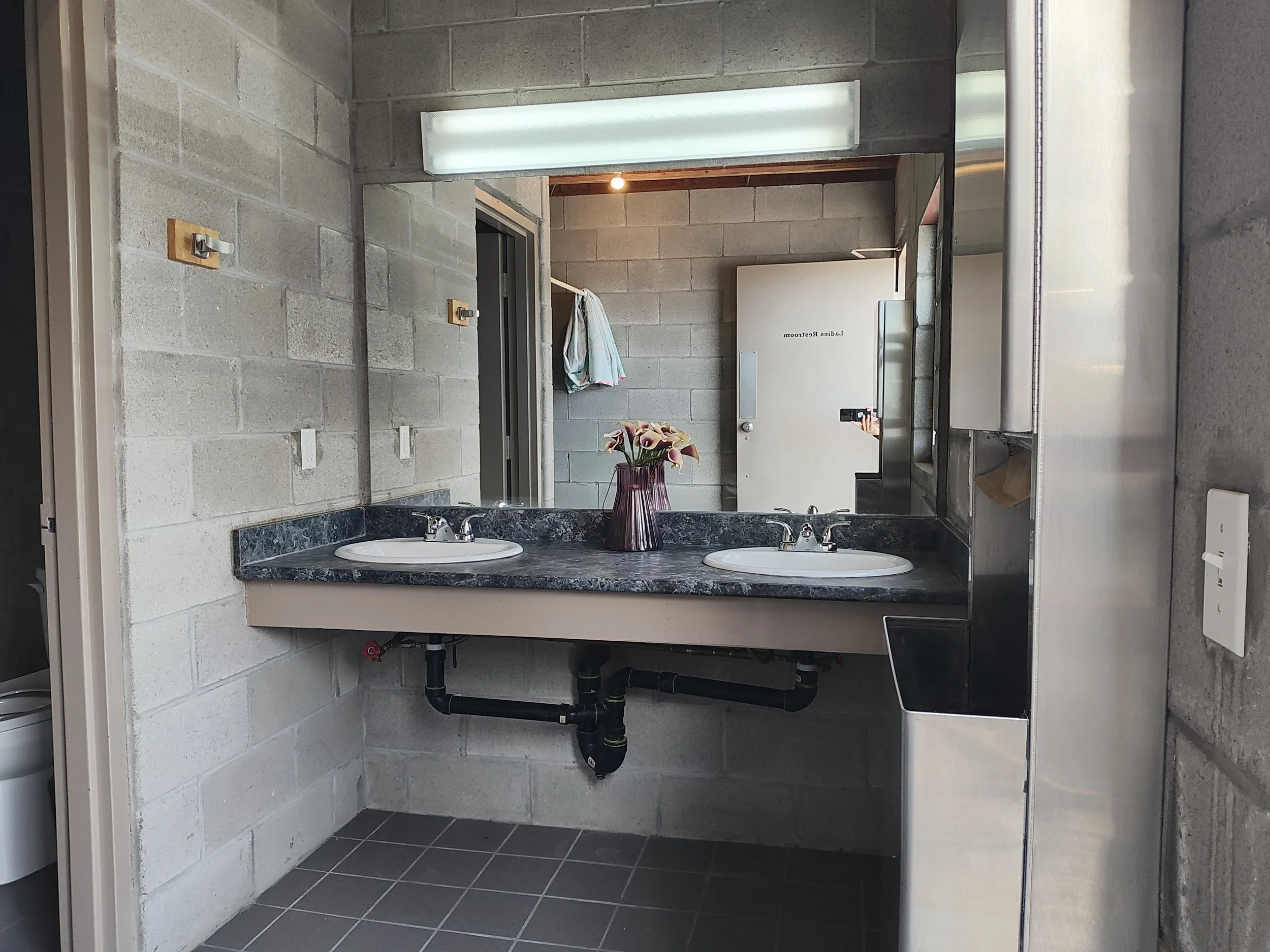 A bathroom mirror reflecting a concrete wall with two hooks, a towel hanging, and a closed door labeled 'Ladies Restroom'. There are two sinks with a granite countertop and a vase with flowers between them. An electric hand dryer is visible on the ri