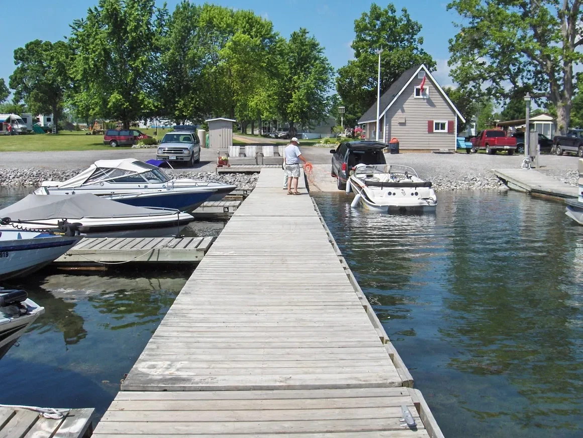 A small marina with boats docked along a wooden pier, a man working on a boat, and a house with trees in the background.