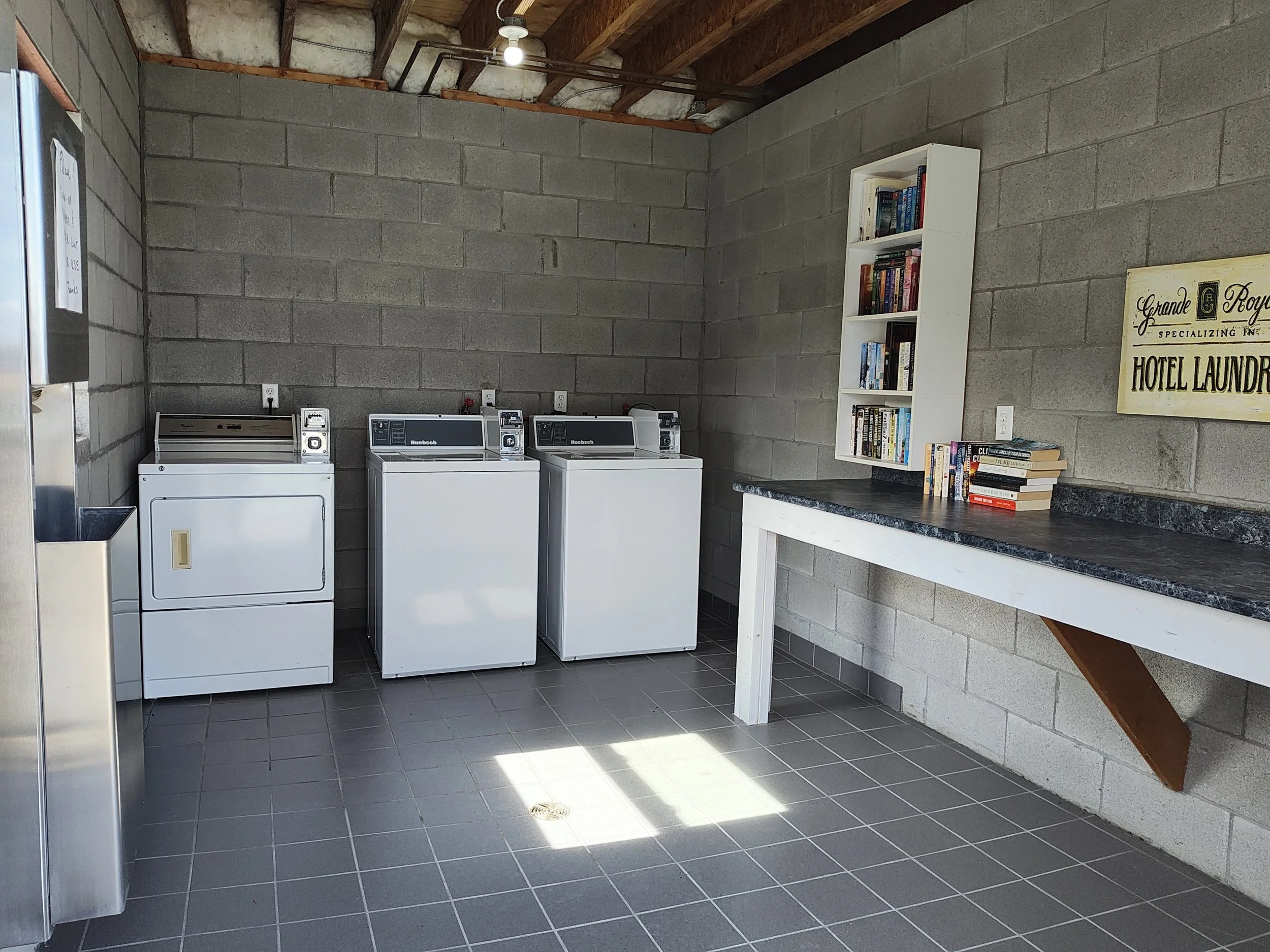 A laundry room with three white washing machines against a gray concrete block wall, a white bookshelf with books, a sign that reads 'Grande Royal', and gray tiled flooring.