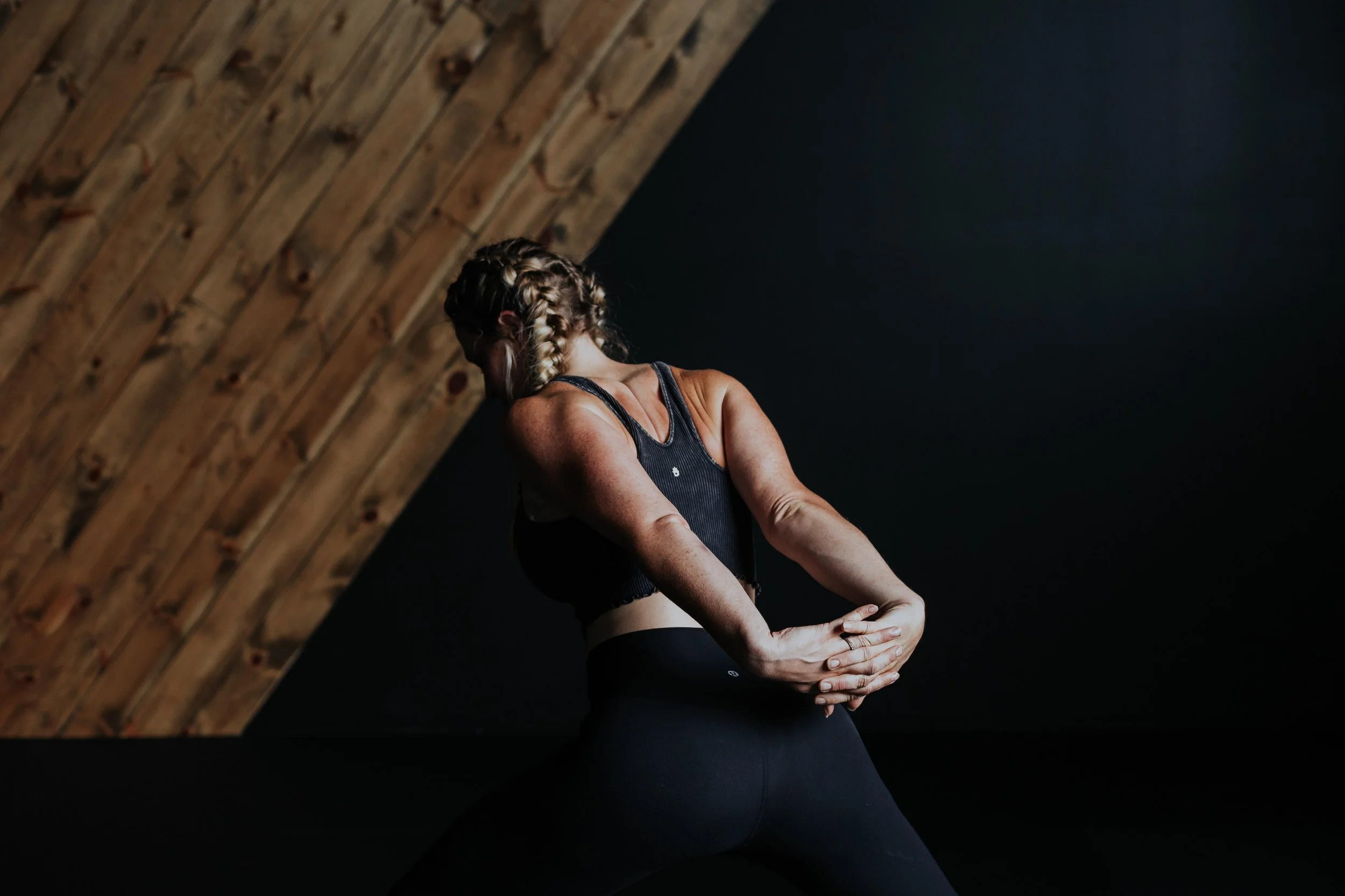 Woman with braided hair practicing yoga or stretching, wearing a black sports top and leggings, indoors with wooden ceiling and dark wall.