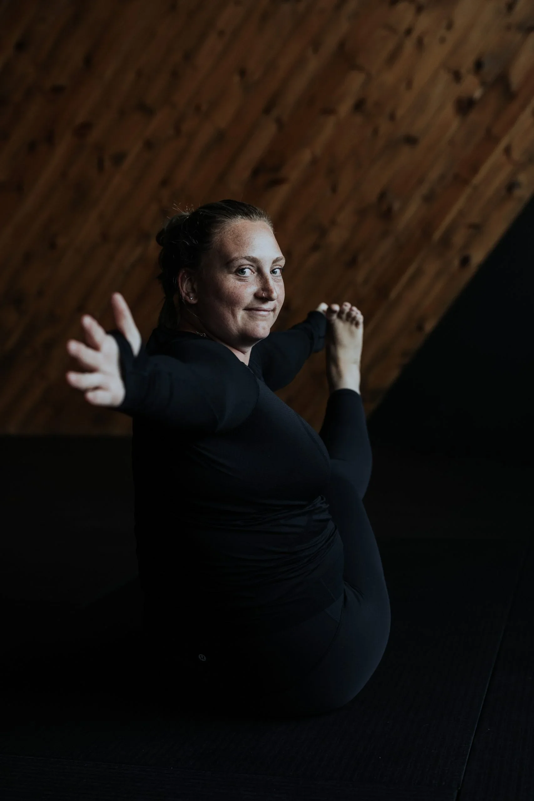 Woman performing a yoga pose on a black mat, with a wooden ceiling background.