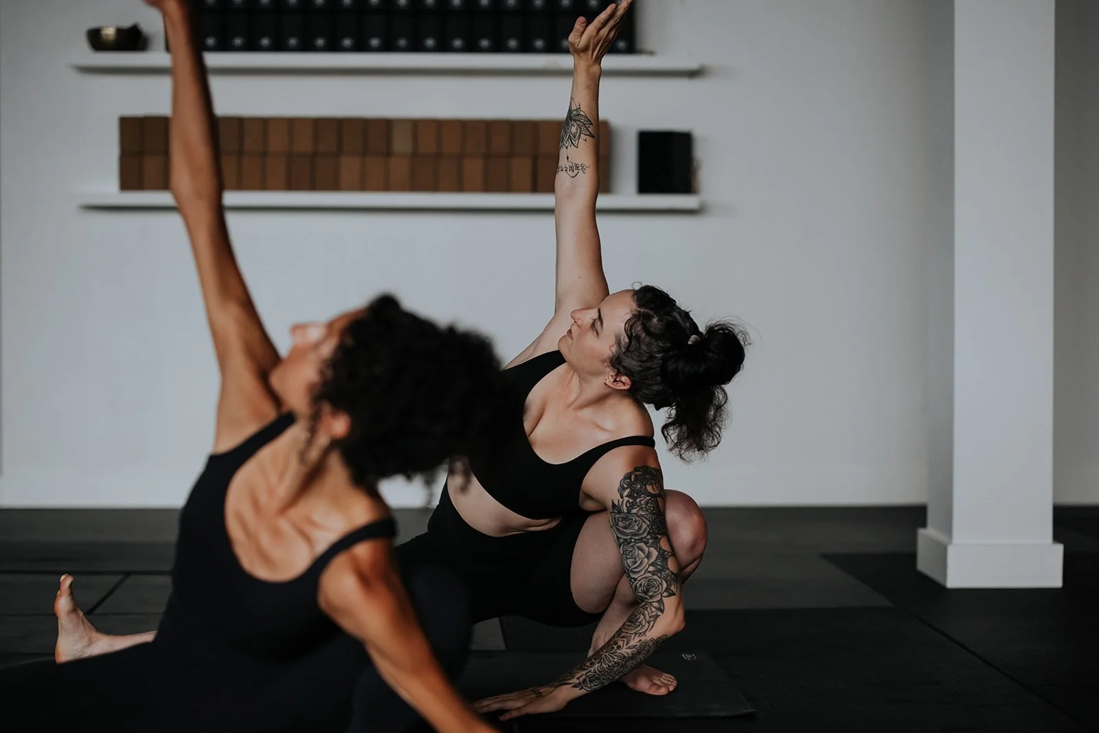 Two women practicing yoga indoors on black mats, with one woman in a black sports bra and the other in black workout clothes, both in a stretching pose. The woman with tattoos is performing a side stretch with her arm extended above her head.