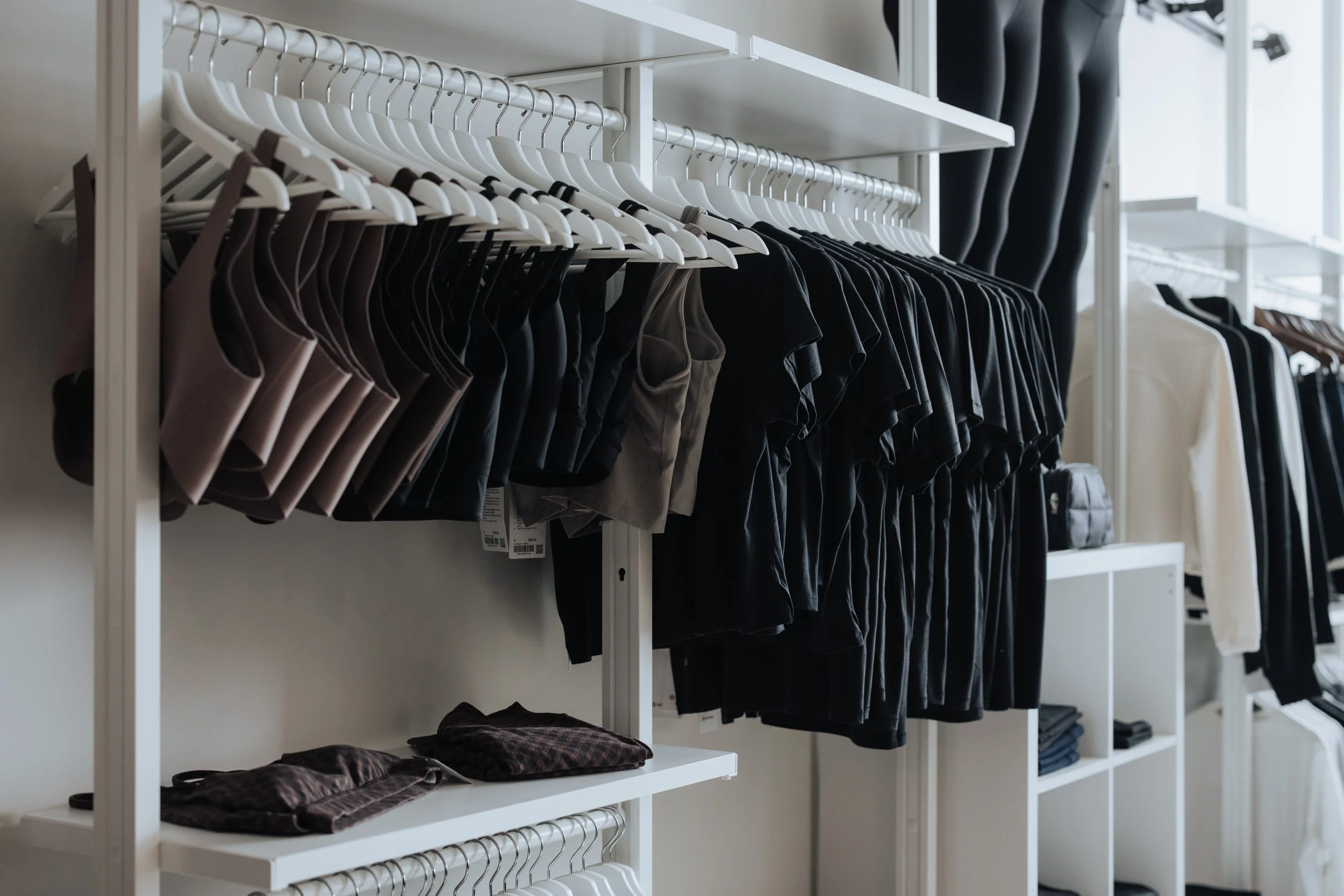 Clothing store display with racks of various clothing items, including black, beige, and white shirts, with folded clothes on white shelves.