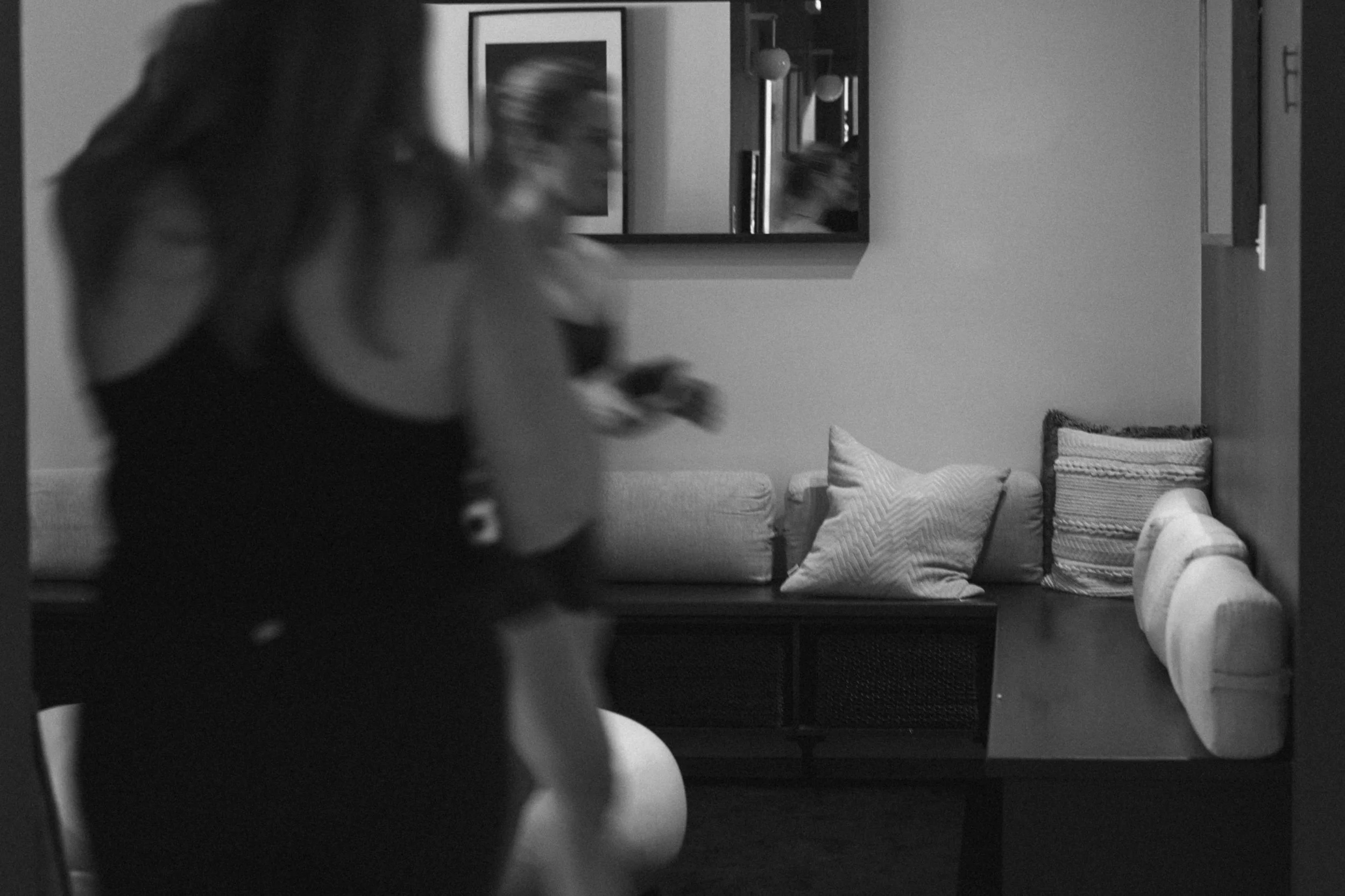 Black and white image of a woman walking past a living room with decorative pillows on a sofa and a framed picture and hanging lights on the wall.