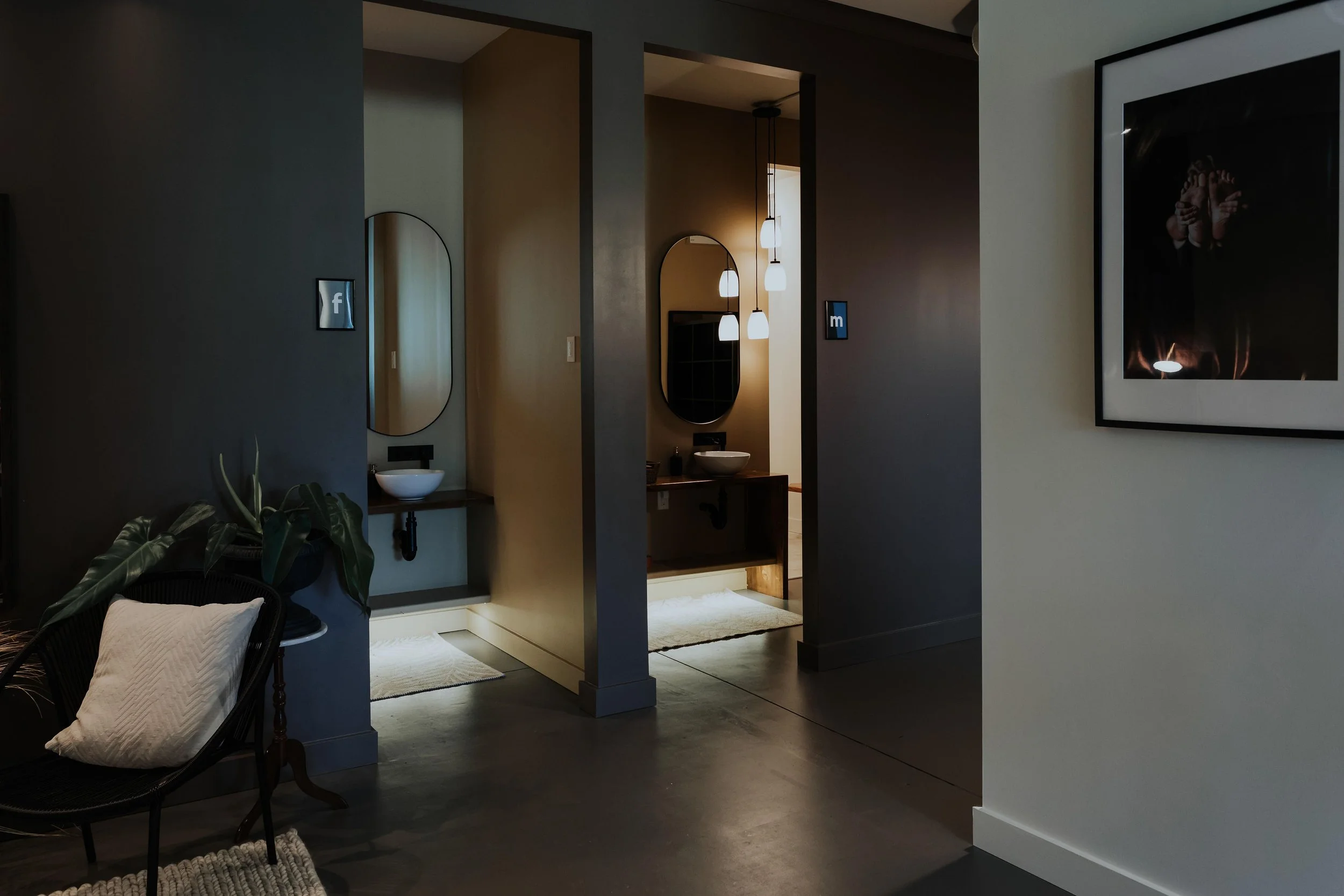 Modern bathroom with a double sink vanity, oval mirrors, pendant lights, and dark-colored walls, adjacent to a cozy sitting area with a plant and a cushioned chair.