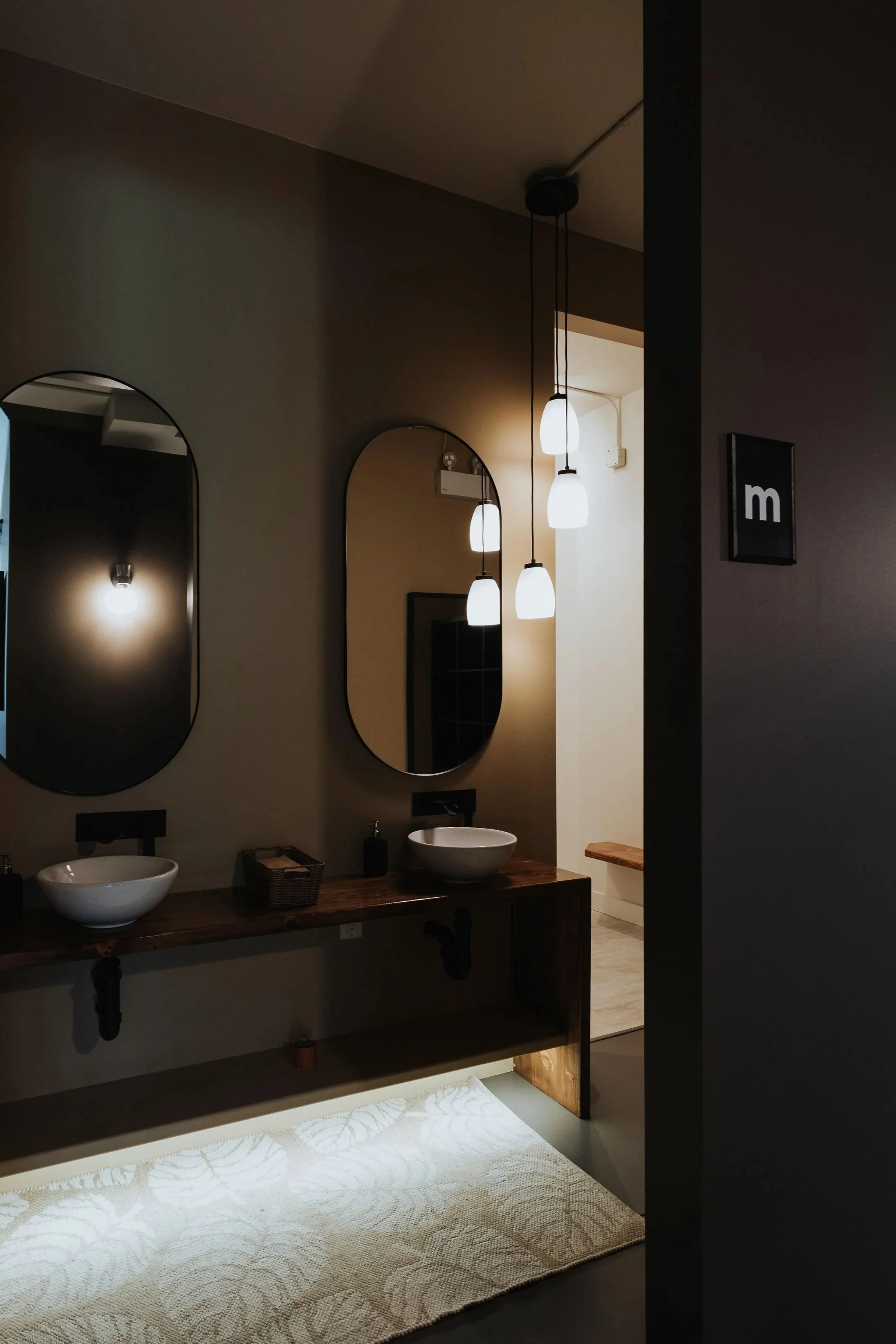 A modern bathroom with two white vessel sinks on a wooden vanity, two oval mirrors, hanging pendant lights, a light-colored rug, and a partial view of a toilet and a wooden bench.