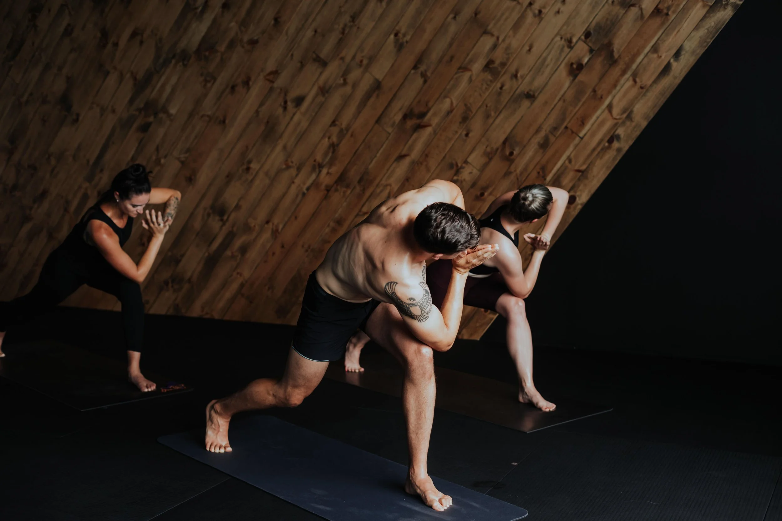 A group of four people, two women and two men, practicing yoga together on black mats in a room with wooden walls. They are in a warrior pose, with legs apart, one knee bent, and arms extended overhead.