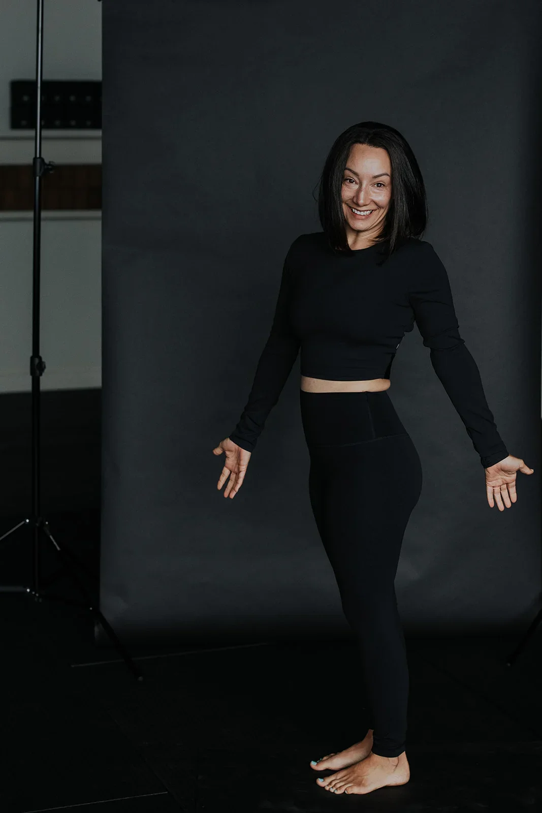 Woman with dark hair in black outfit standing barefoot in front of a black backdrop, smiling.