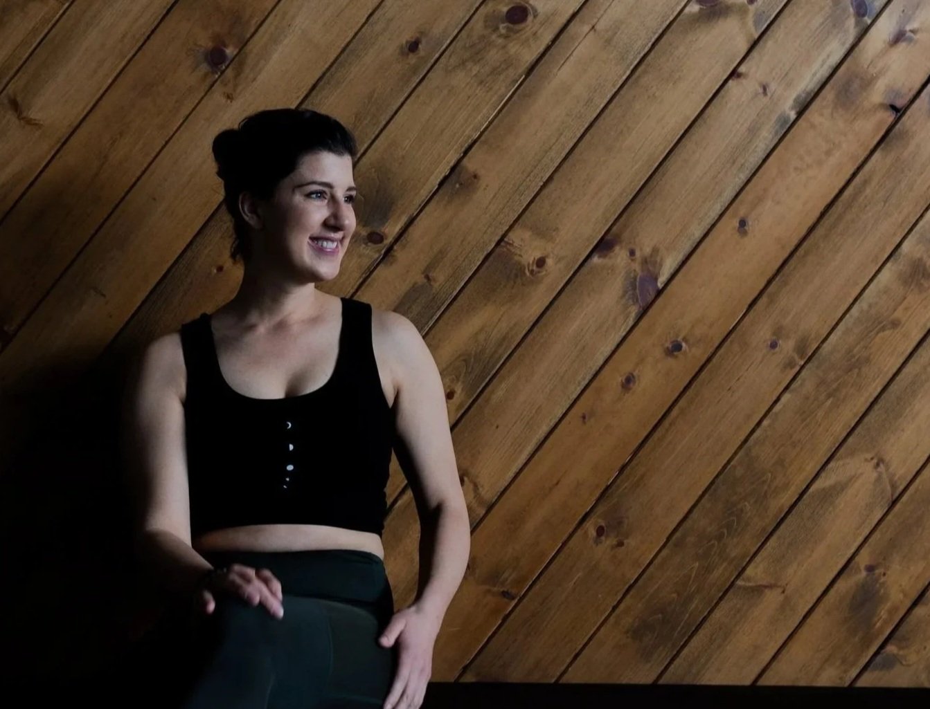 A woman with short dark hair smiling, standing against a wooden wall, wearing a black sleeveless crop top and high-waisted dark pants.
