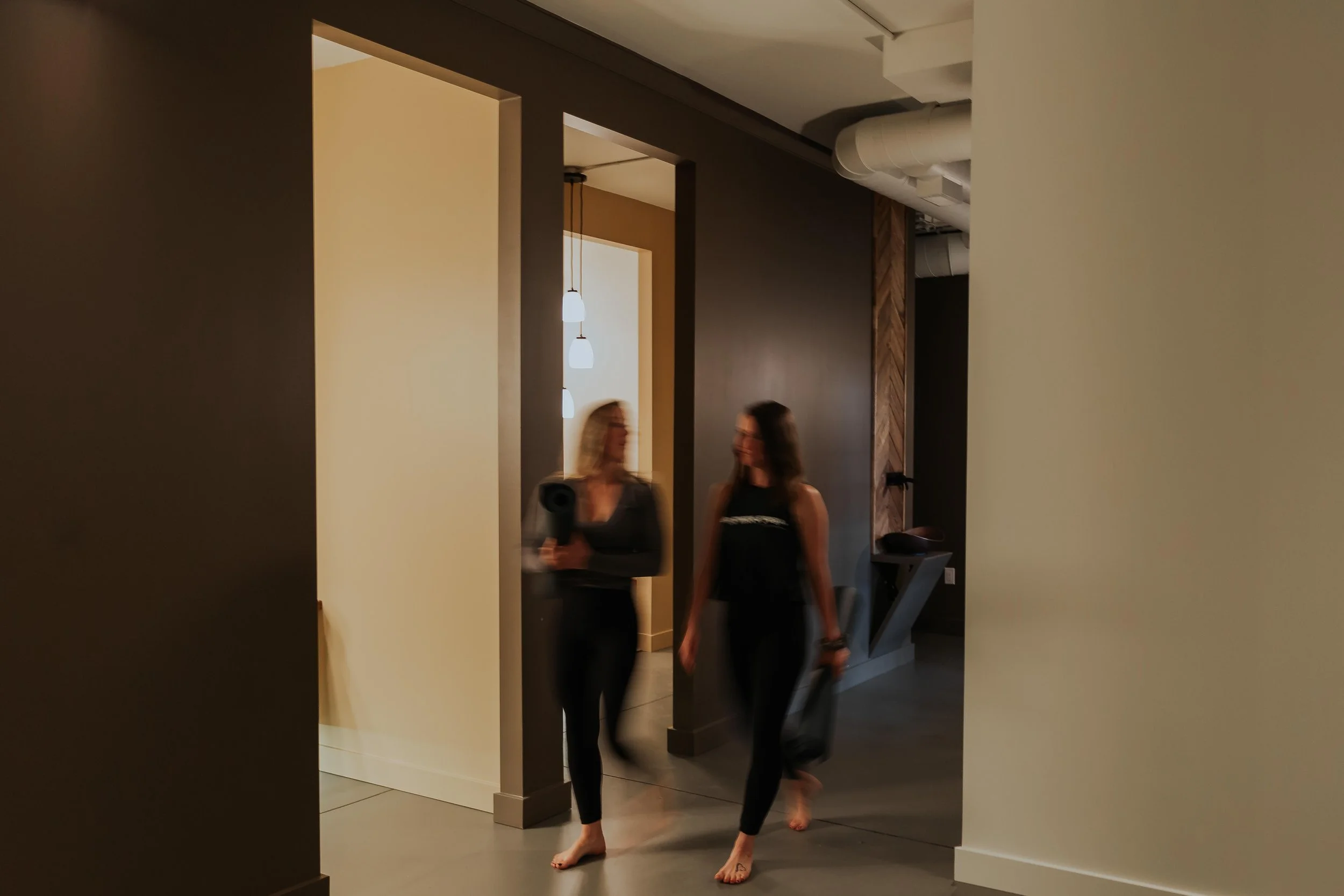 Two women walking barefoot in a modern interior space with warm lighting, dark walls, and a white wall with hanging lights in the background.