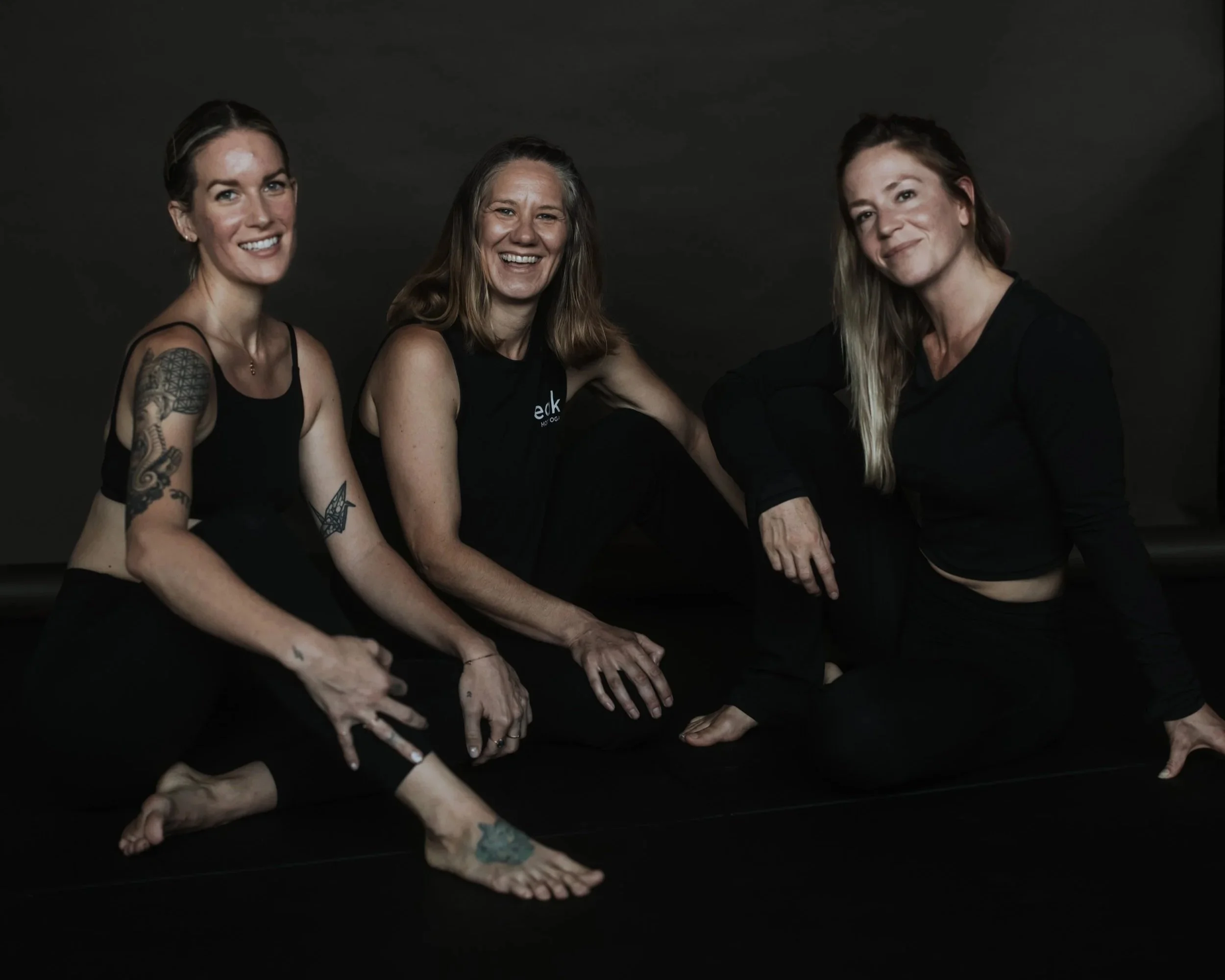 Three women sitting on the floor against a black background. They are smiling and looking at the camera. The woman on the left has tattoos on her arm and leg, and is wearing a black tank top. The woman in the middle has long brown hair, and is wearing a sleeveless black top. The woman on the right has long blonde hair and is wearing a black top. Their hands are resting on the floor in front of them.
