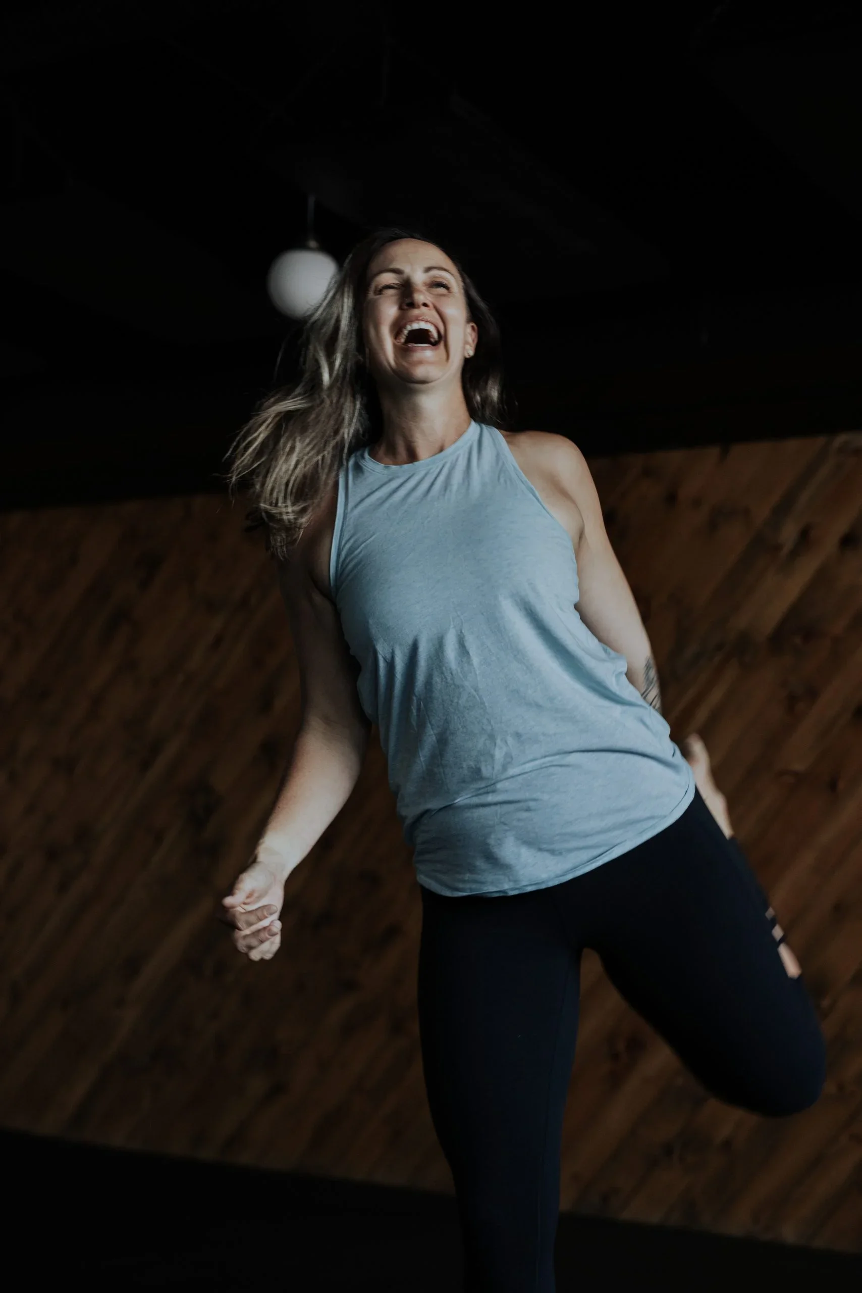 A woman with long hair wearing a light blue tank top and black pants is smiling while holding hands with someone. The setting appears to be indoors with a dark ceiling and wood-paneled wall.