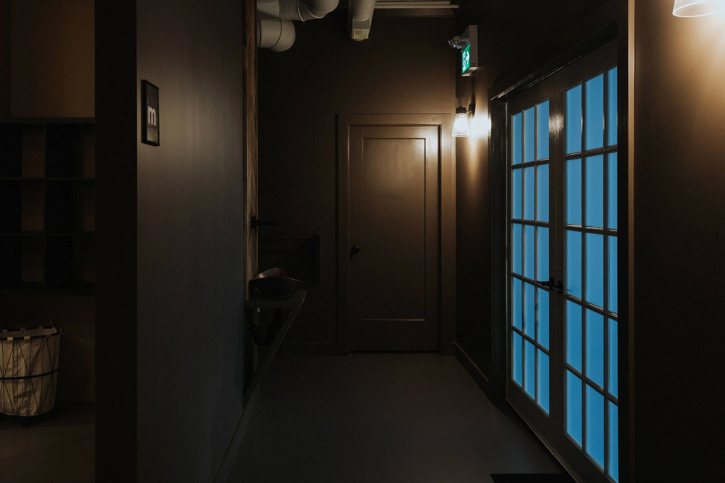Dimly lit interior with a glass-paneled door on the right, visible outside is a blue sky or sunset, with a wall-mounted emergency exit sign above the door, and a window reflecting the outdoor scene. The room has a dark staircase, a vinyl basket with a wire base, and a small table with an object on it.