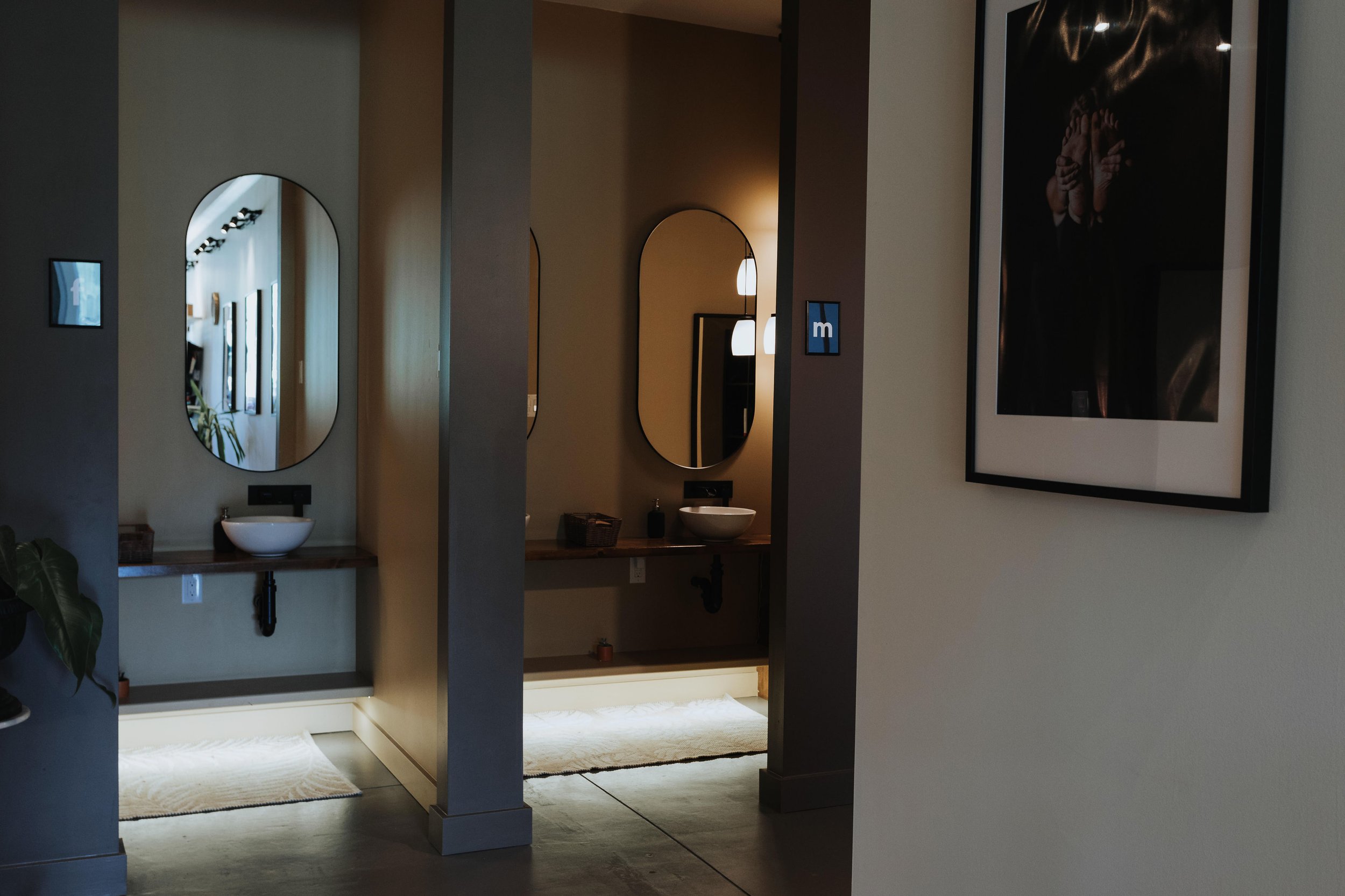 A modern bathroom with two oval mirrors above sinks, a framed photo on the right wall, and a well-lit space with pendant lights visible through an archway.
