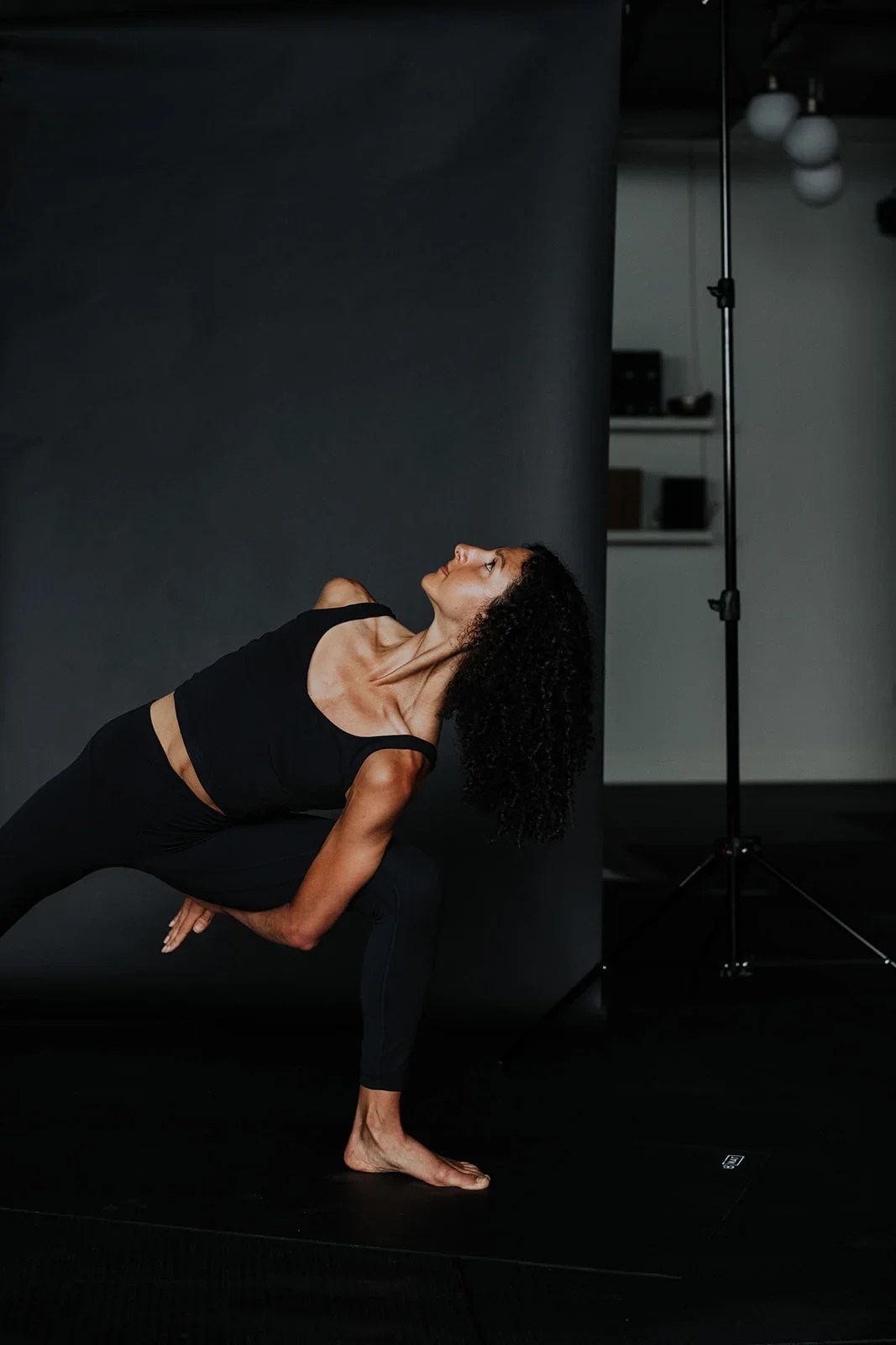Woman with curly hair practicing yoga in a studio, balancing on one leg with her head tilted back and arms crossed over her body, against a dark background with studio equipment visible.