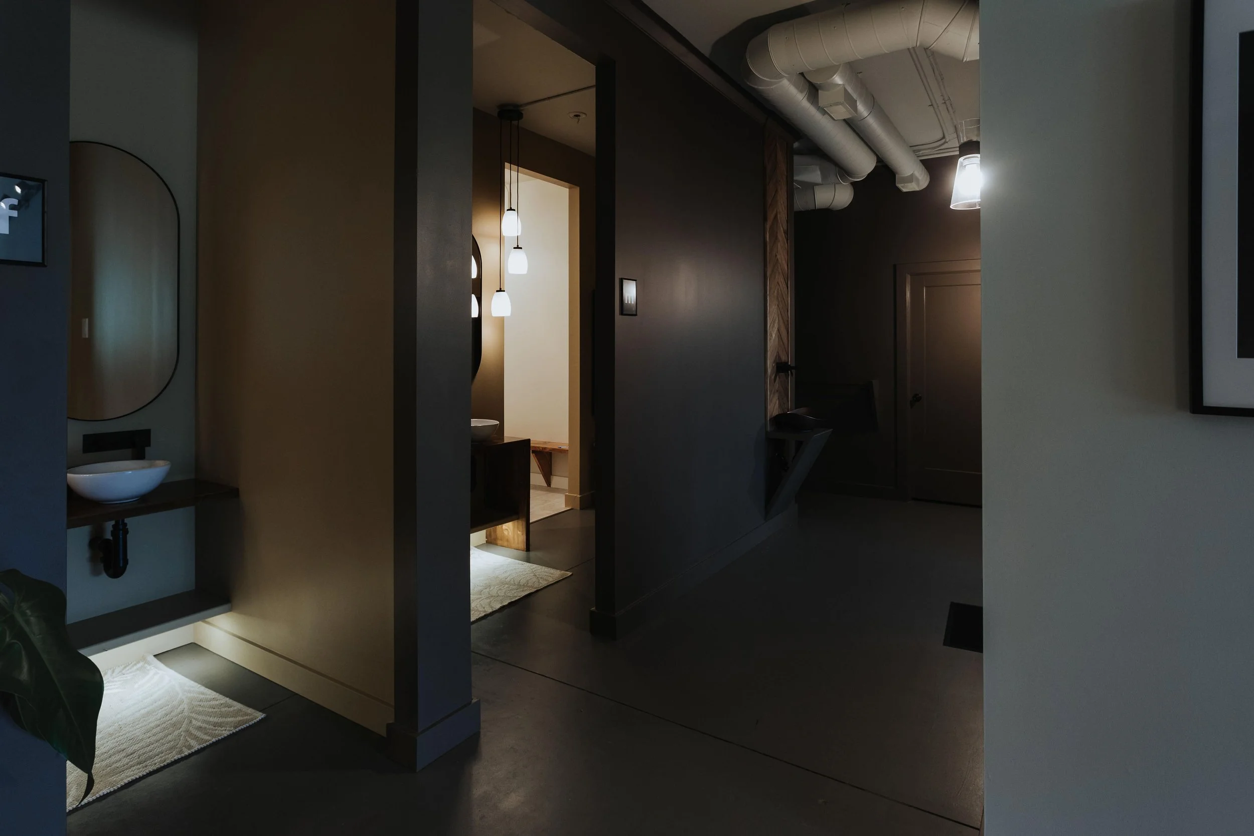 Interior view of a modern apartment with dark walls, a hallway, and visible ceiling ducts. There are hanging pendant lights illuminating a space, with a bathroom or vanity area visible in the background. A mirror and a vessel sink are also visible.