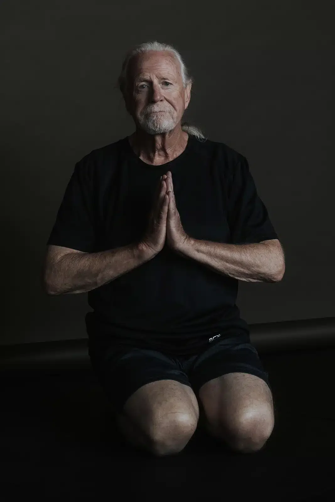 An elderly man with gray hair and a beard is kneeling with his hands pressed together in a prayer position, wearing a black T-shirt and shorts, against a dark background.