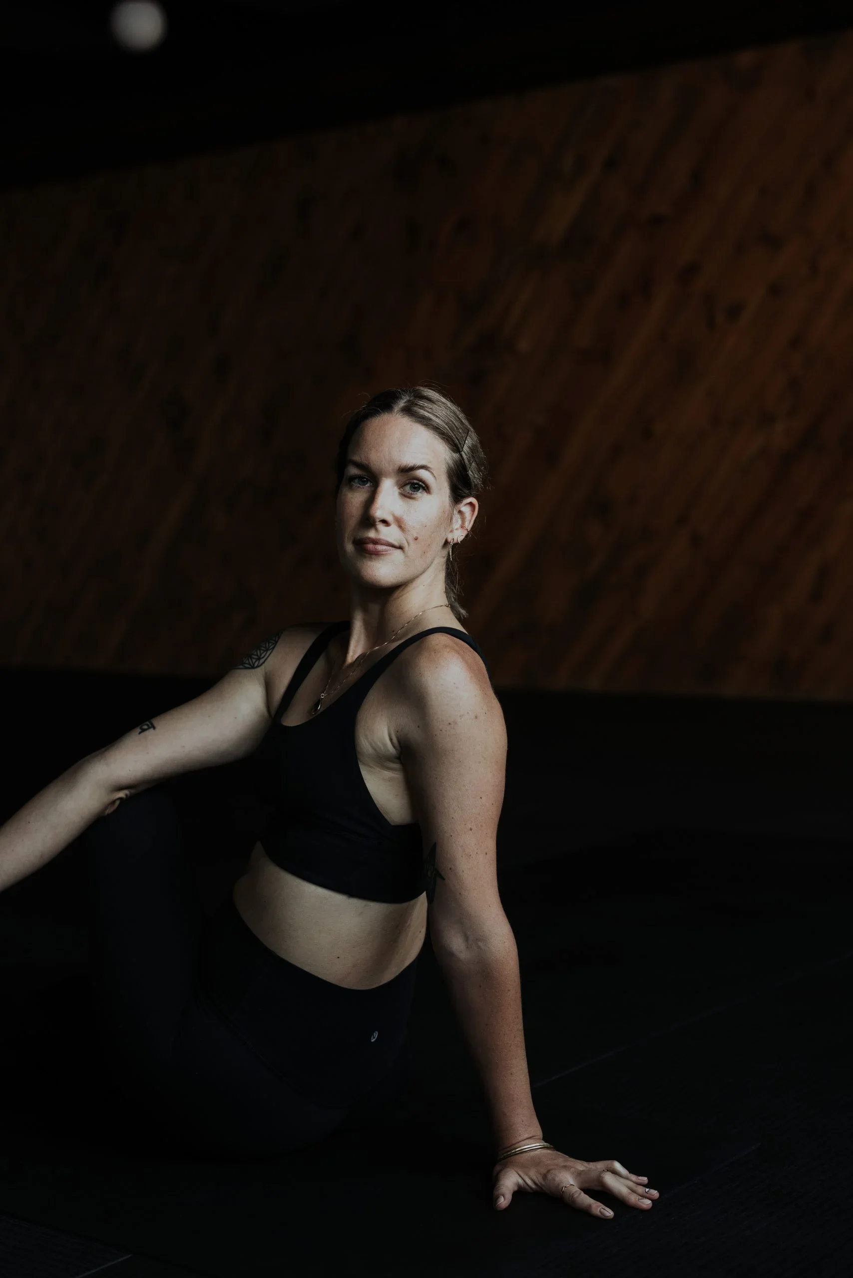A woman in black athletic clothing stretching or exercising indoors against a brown wooden wall.