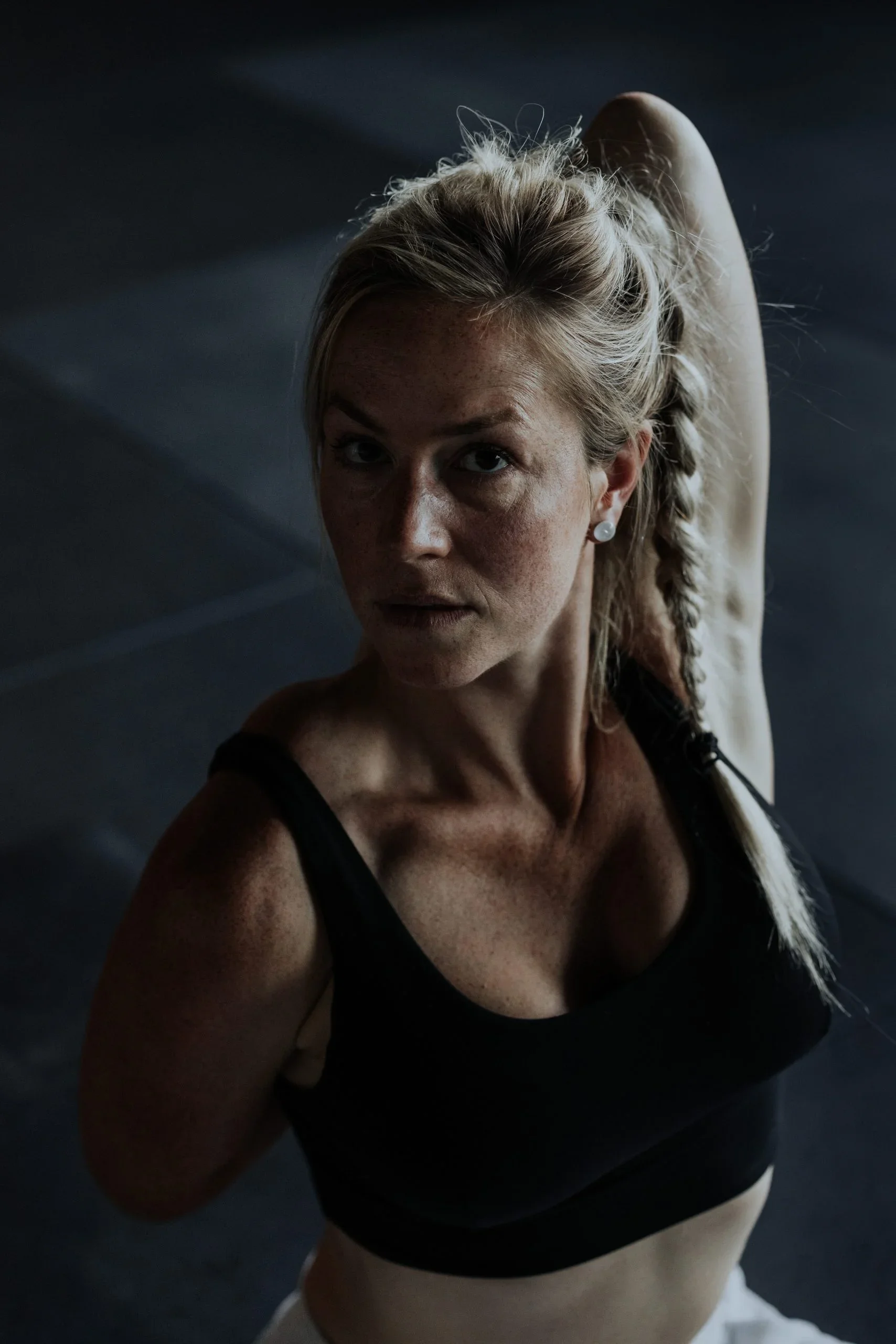 A woman in workout attire performing a stretching exercise in a dark gym, with her arm raised above her head and her hair in a braid.