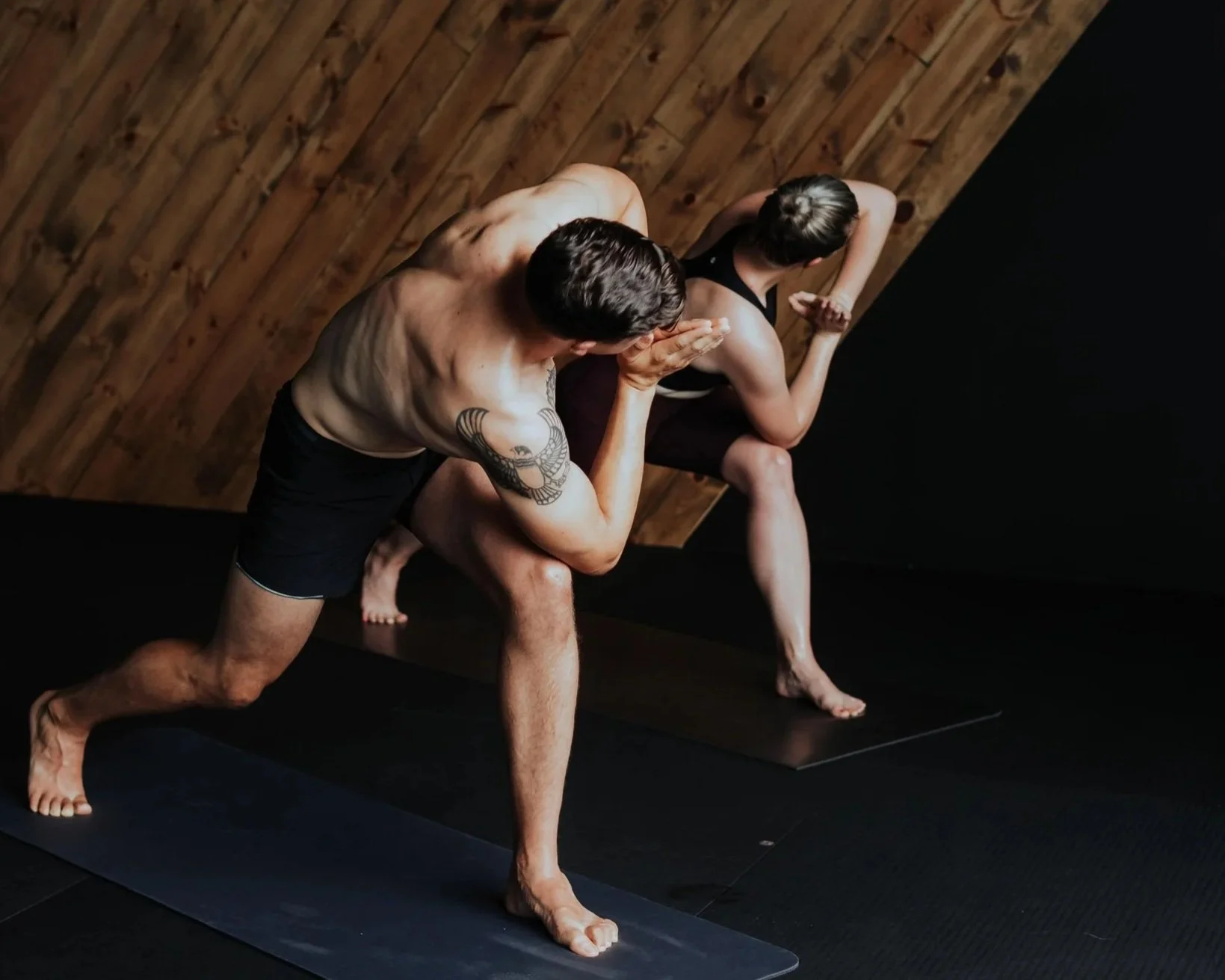 A man and a woman practicing yoga together on black mats in a room with wooden walls. The man is shirtless, wearing black shorts, and has a tattoo on his arm. He is in a lunge position with one arm extended and the other hand near his face. The woman is wearing a black sports bra and shorts, with her hair tied back. She is in a side stretch pose with one arm reaching over her head and the other hand on the ground.