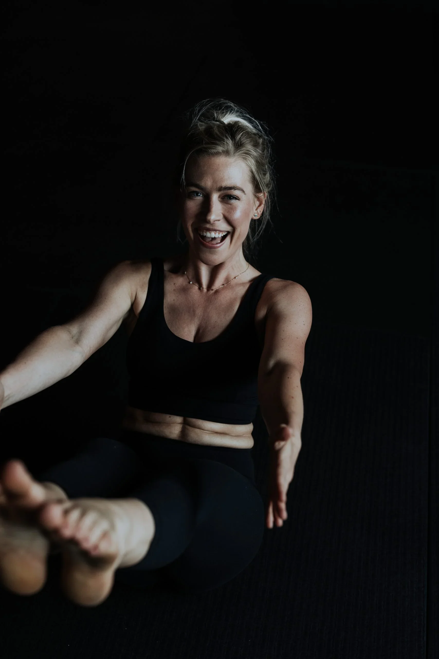A woman in black workout clothing taking a selfie while practicing yoga on a black mat. She is sitting cross-legged with her hands extended forward and smiling at the camera.
