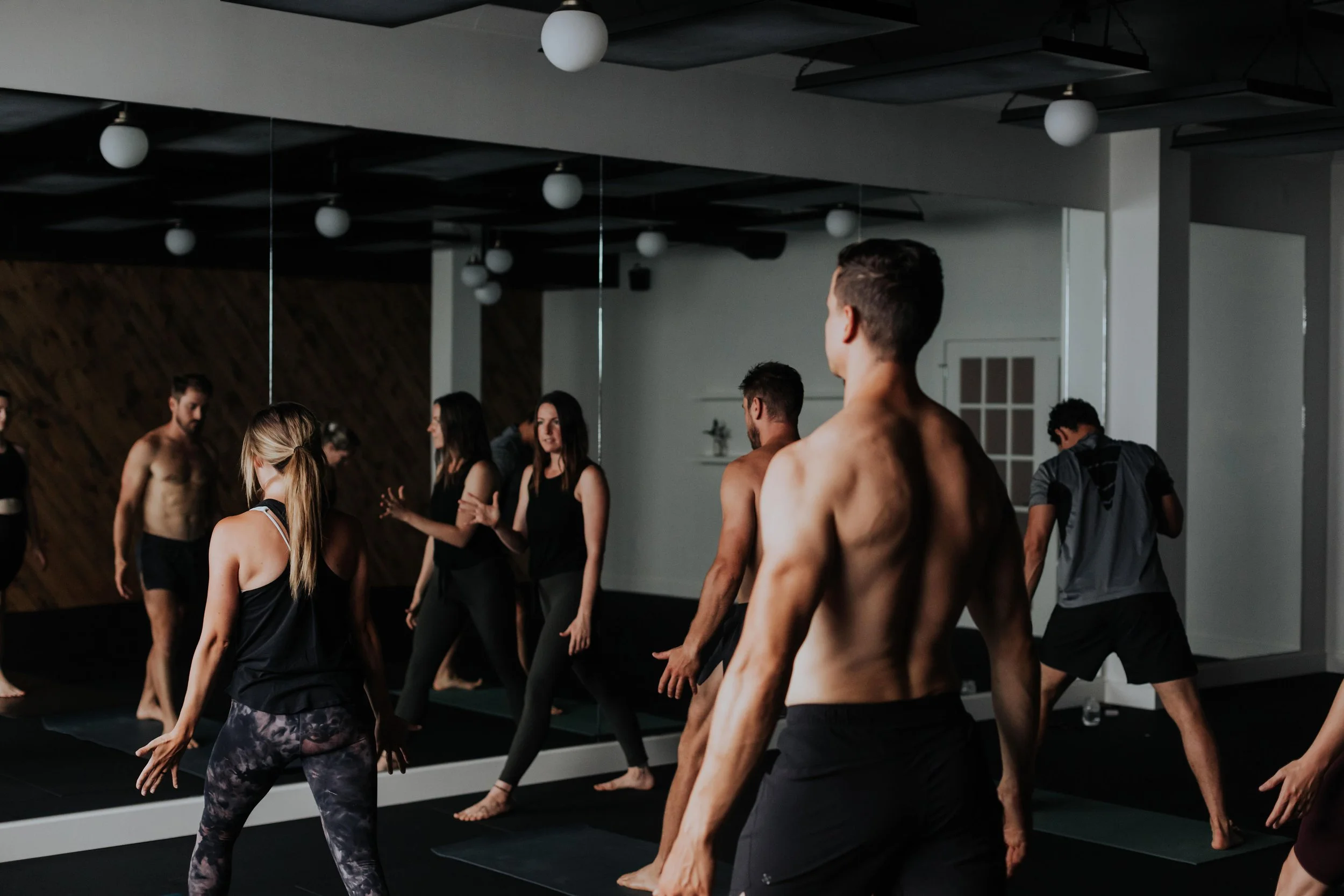 Group of people participating in a yoga or stretching class in a studio with dark walls and ceiling, and round lighting fixtures.