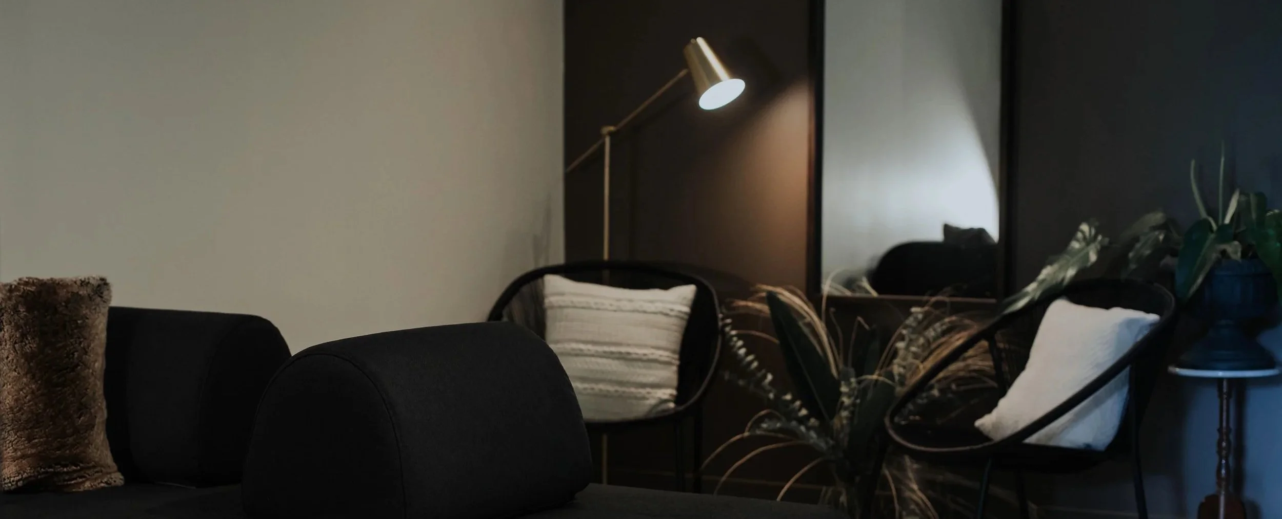 A cozy living room corner with black chairs, a cushion, a wall lamp, and a large mirror reflecting part of the room, including a plant with broad leaves and a white cushion.