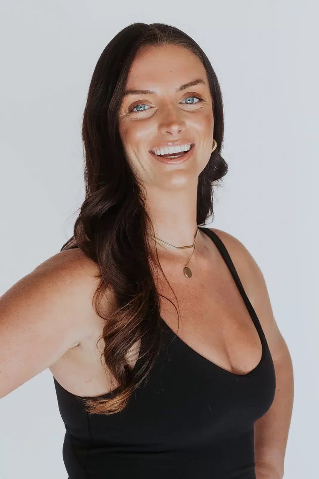 Portrait of a smiling woman with long dark hair, blue eyes, wearing a black tank top and layered necklaces, set against a white background.