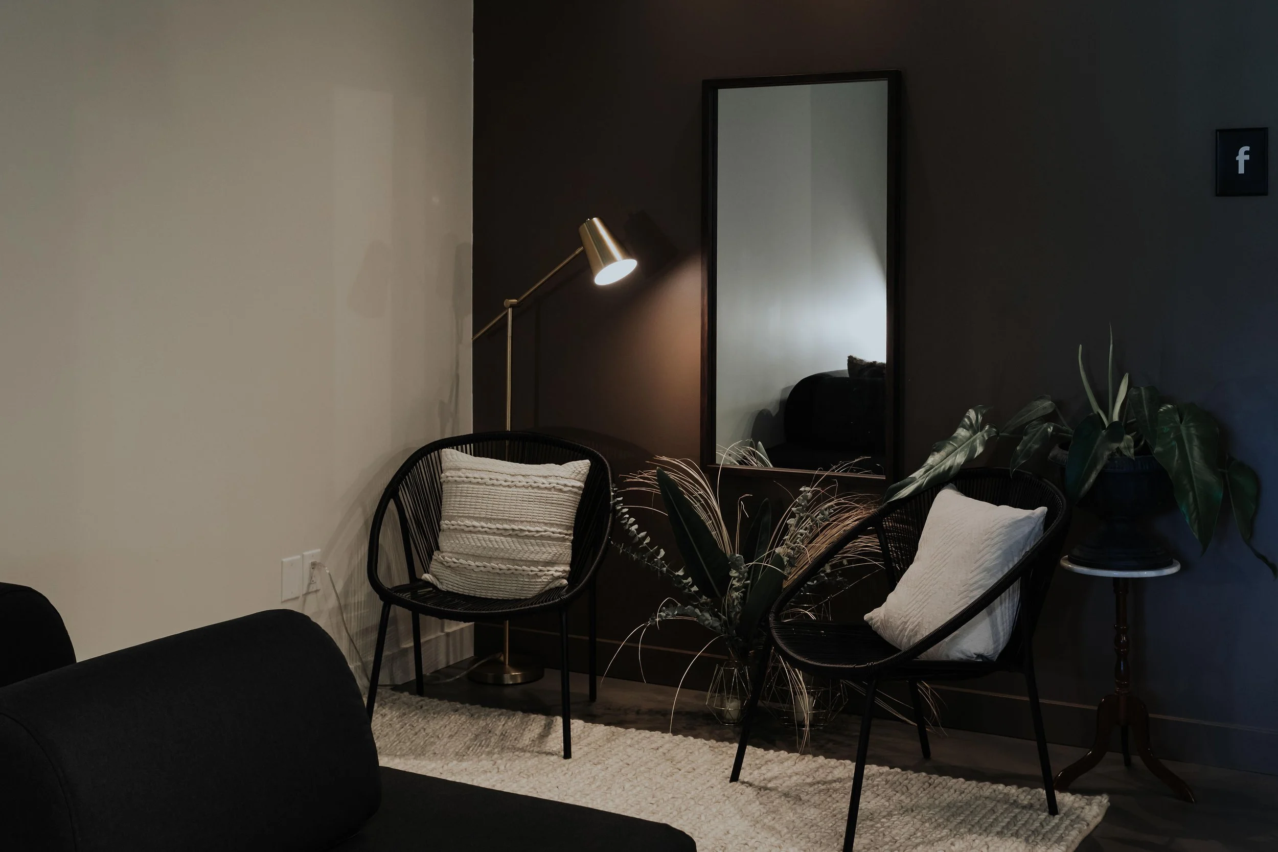 A cozy living room corner with a black reclining chair, a round side table with a plant, a full-length mirror, a standing lamp, and framed Facebook logo on the wall.