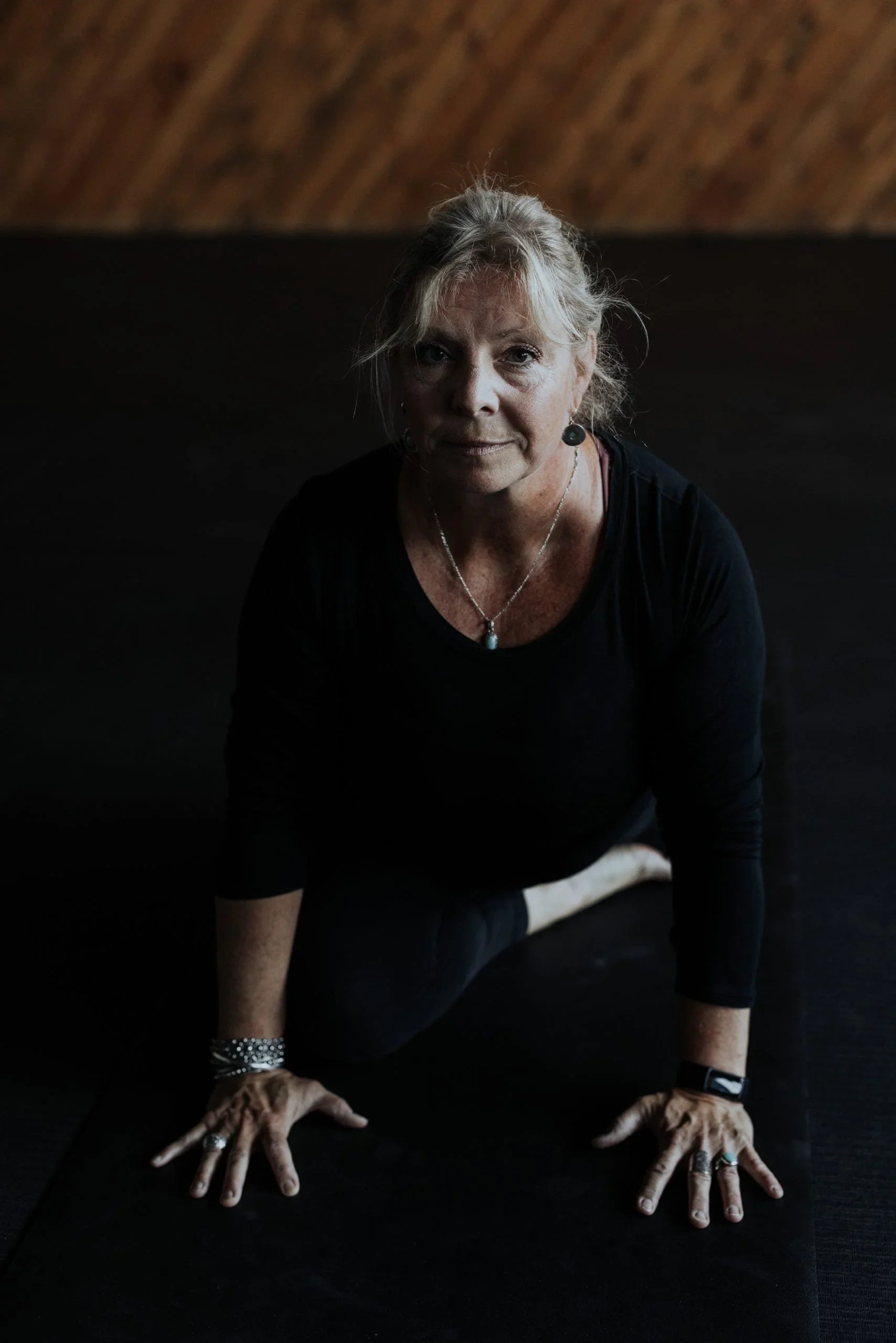 A woman with gray hair and jewelry, dressed in black, is performing a yoga pose on a black mat. She is positioned on her hands and knees with her arms extended. The background features a wooden wall.