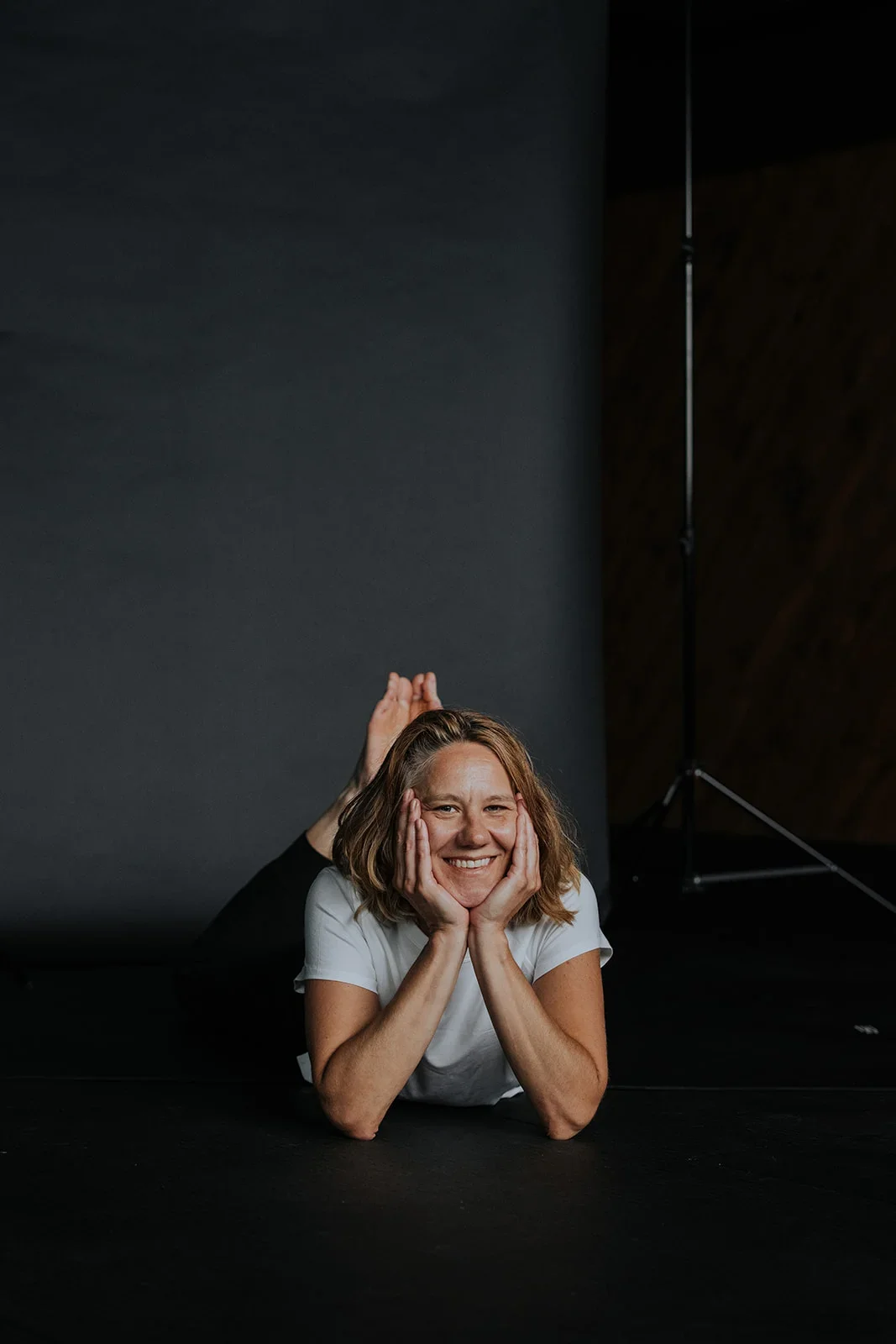 Woman smiling in a yoga pose lying on her stomach with her face resting in her hands on a black mat, dark background.