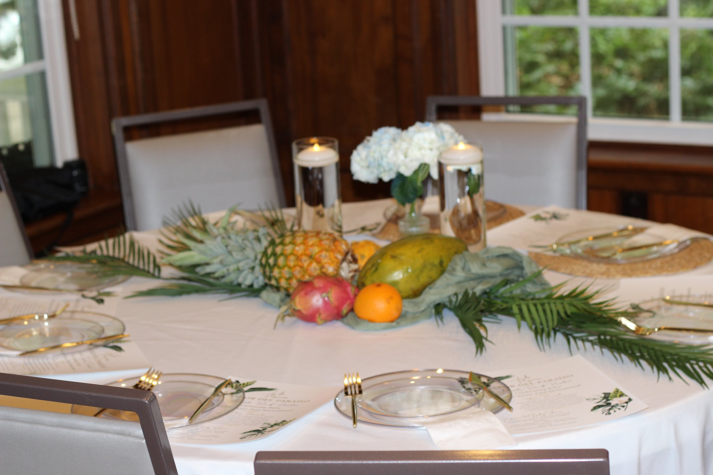 Custom elegant baby shower designed by MPR Events LLC, with tropical leaves, tropical fruit, florals and candlelight centerpieces, set for a meal with elegant plates, gold utensils, and glassware, in a reserved space at Dorothy B. Oven Park.